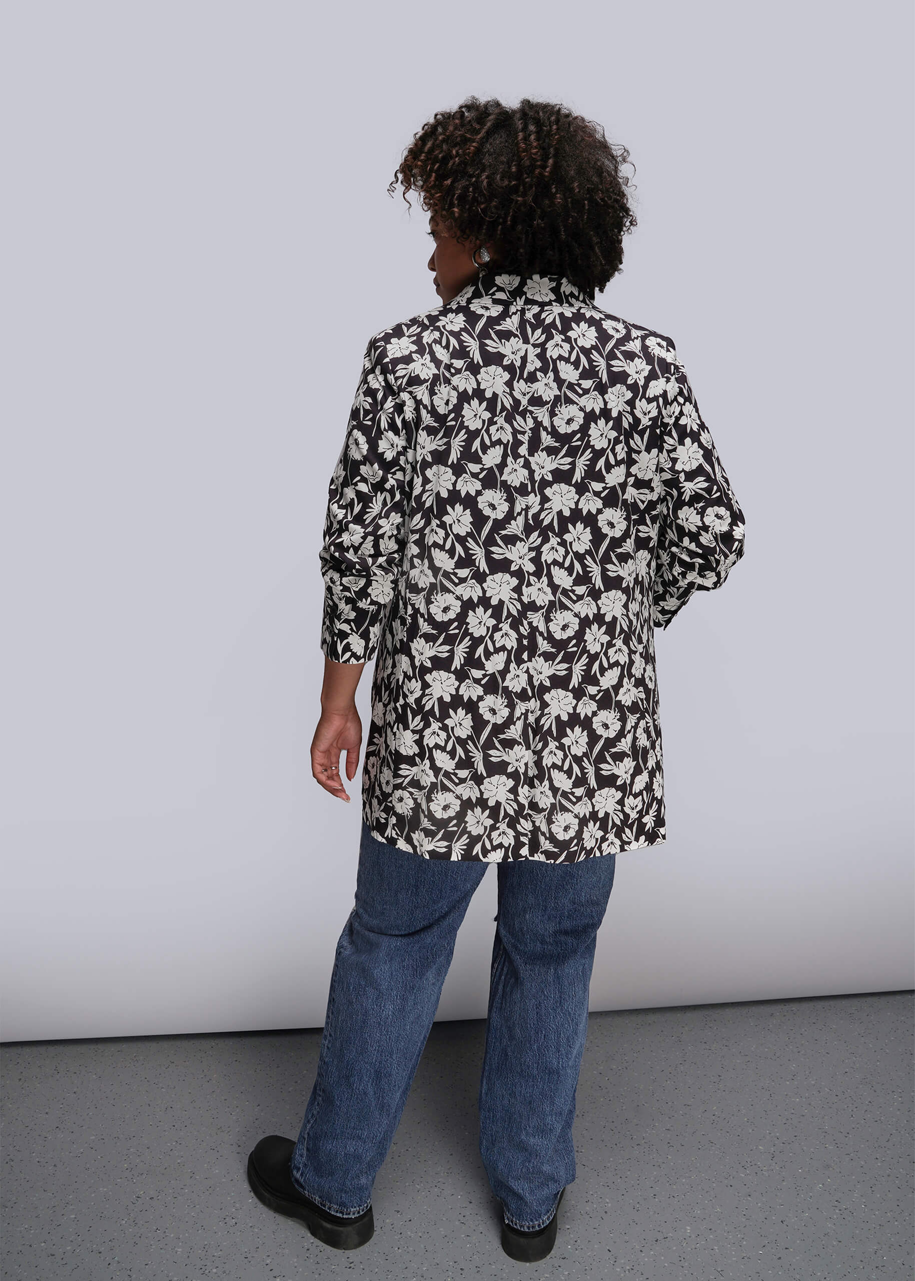 1X Back of person wearing a black and white floral blazer and blue jeans against a plain background