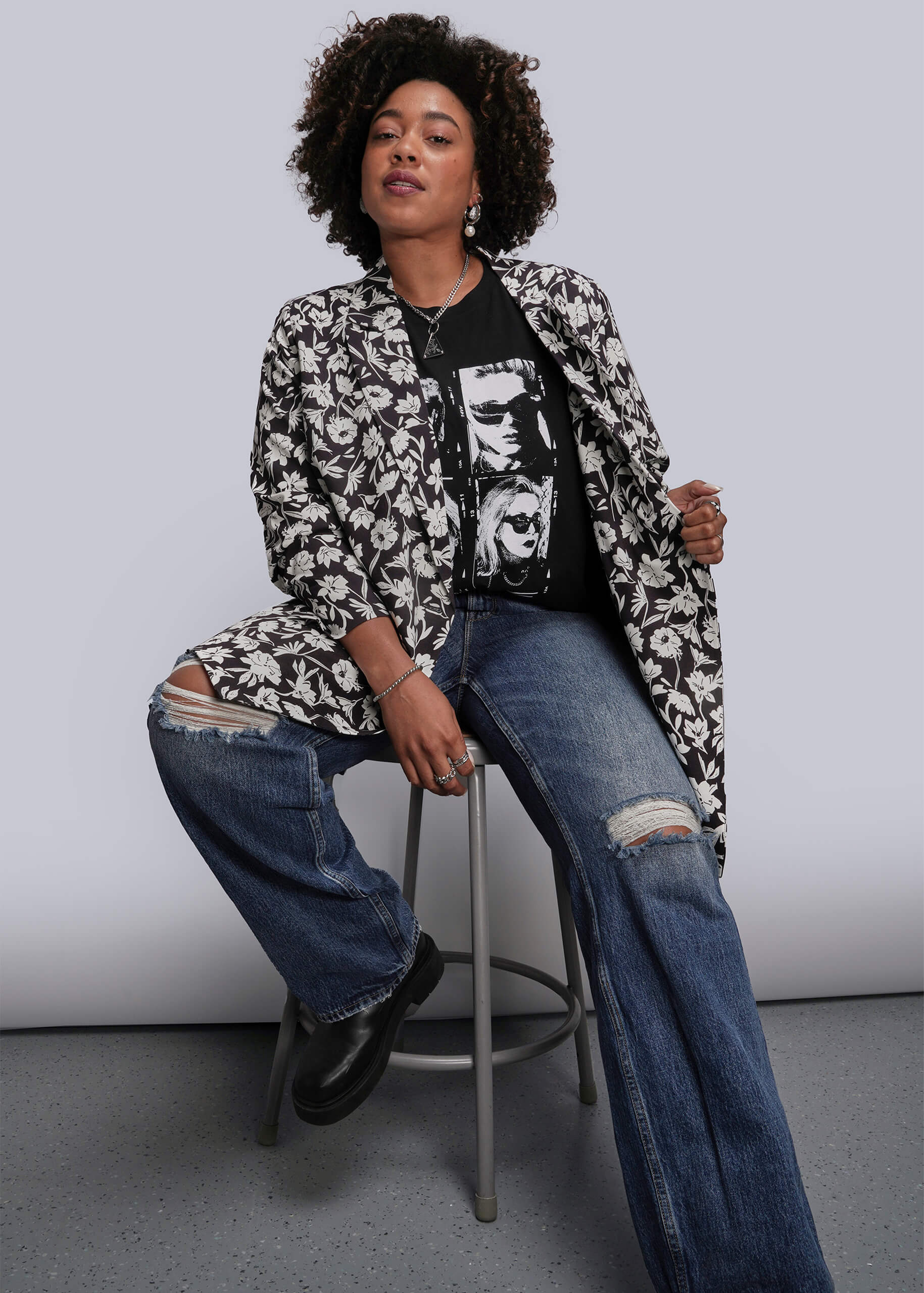 1X=Person sitting on a stool wearing a floral blazer, graphic t-shirt, and jeans against a plain background