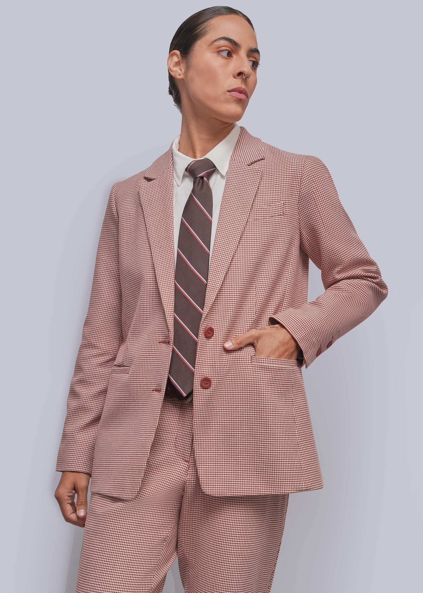 A model wearing a cinnamon and white micro-check suit, including a blazer and trousers, styled with a white collared shirt and a brown and pink striped tie, against a solid gray background.