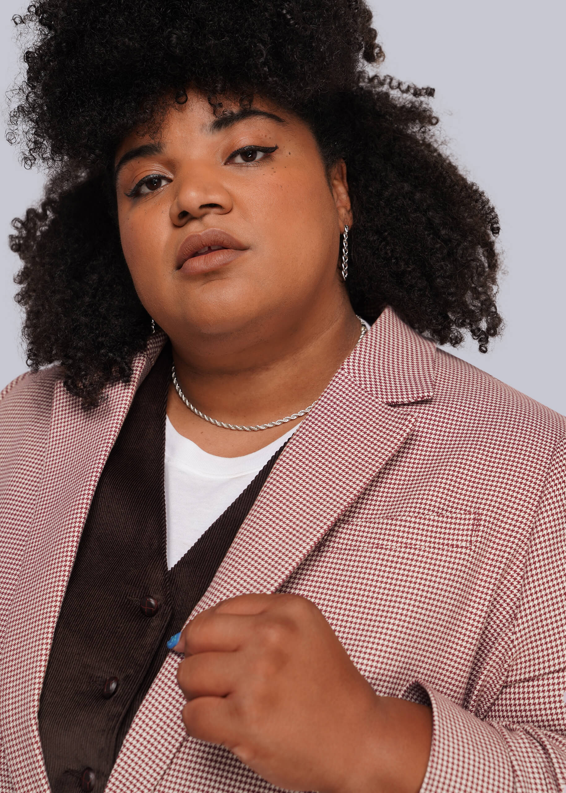 1X=A close-up portrait of a person with dark curly hair wearing a red and white micro-check blazer over a dark brown vest and white t-shirt, against a light gray background.