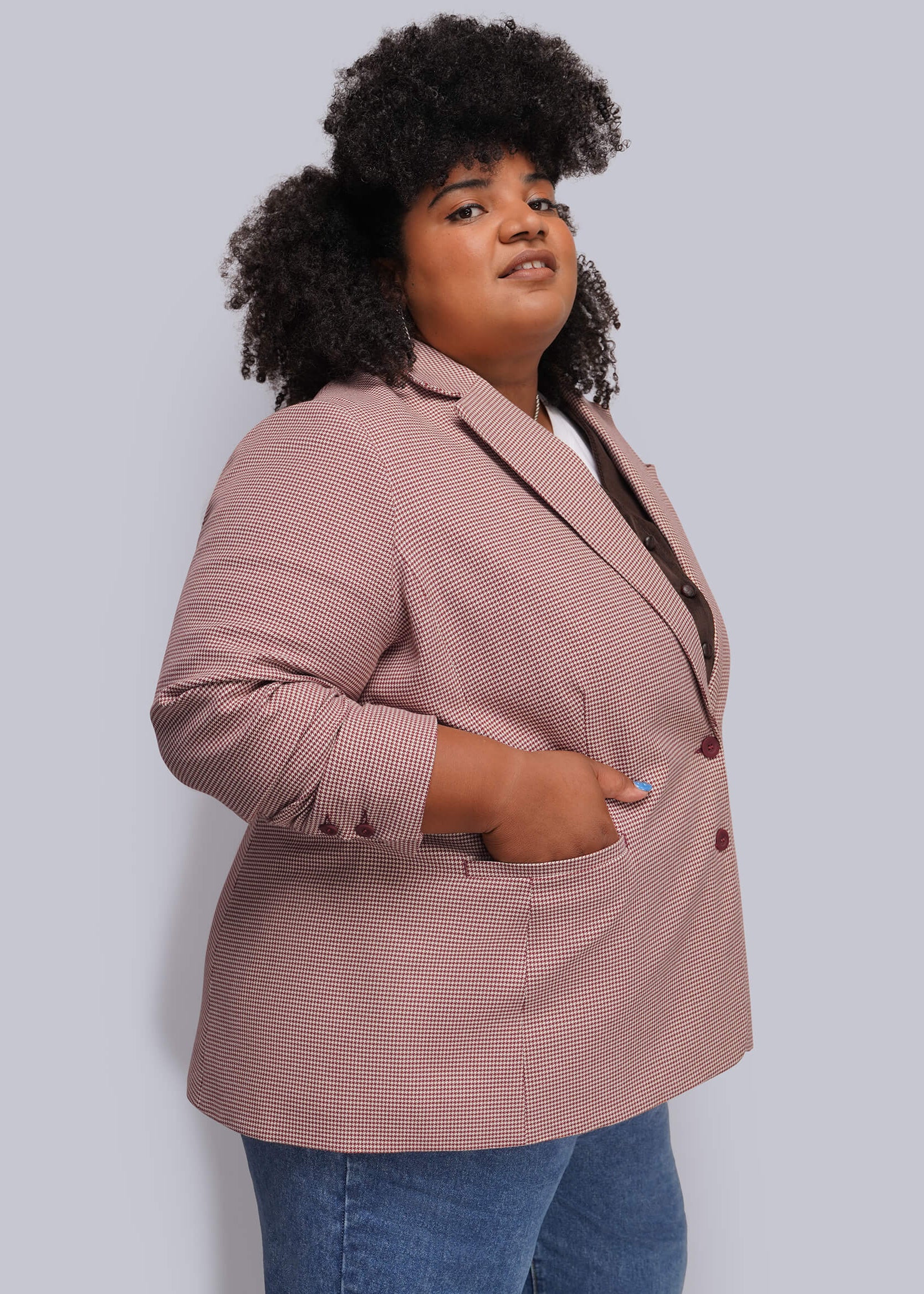 1X=A woman wearing a dusty red houndstooth blazer with blue jeans, standing against a solid light gray background.