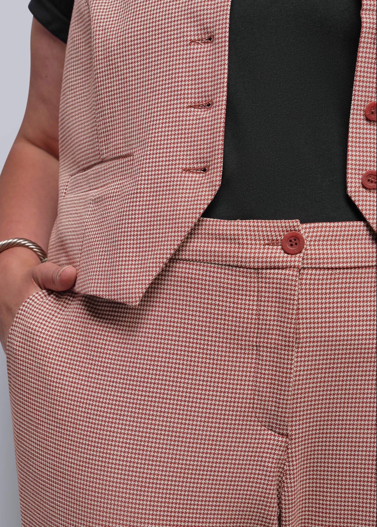 1X=A close-up of a person wearing a matching two-piece suit set with a fine red and beige houndstooth pattern, featuring a buttoned vest and trousers, styled over a black top.