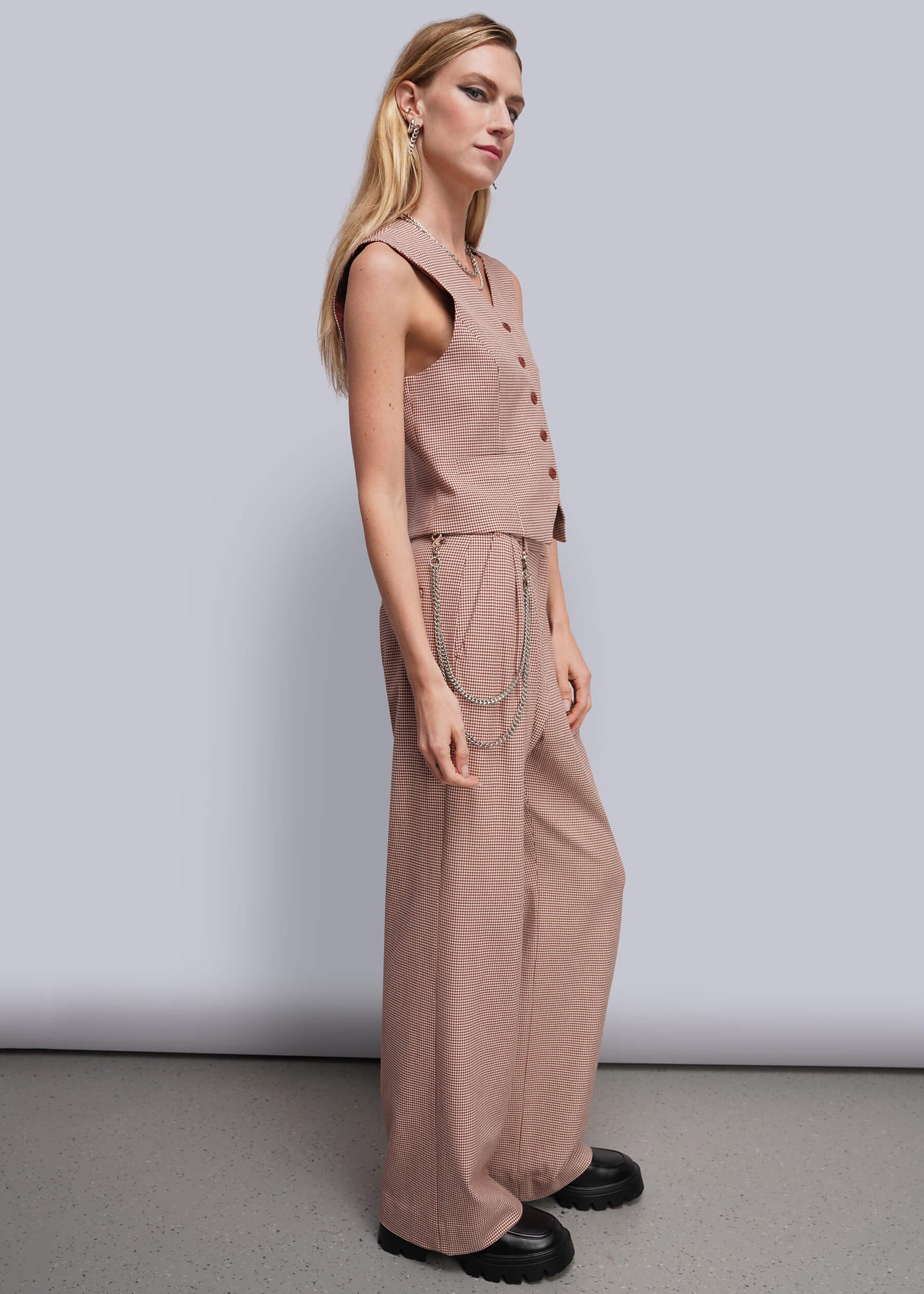 A woman modeling a sleeveless dusty red suit with wide-leg pants, paired with black platform shoes against a light gray studio background.