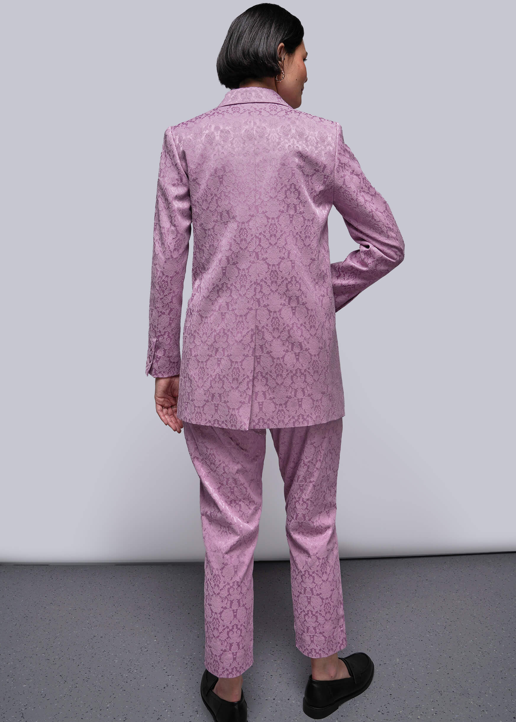 S=Person wearing a pink patterned suit standing against a plain background