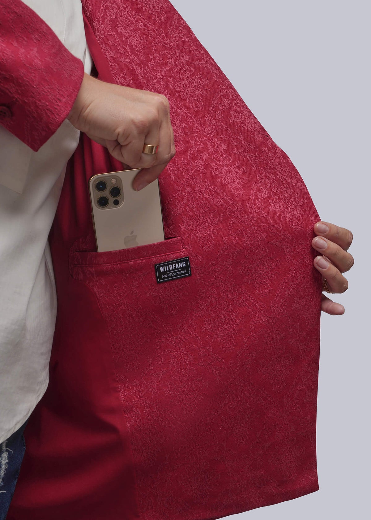 1X=A close-up of a person wearing a red textured blazer and holding a gold smartphone in the front pocket of the blazer. A small black brand label reading 'WILDFANG' is visible on the pocket.