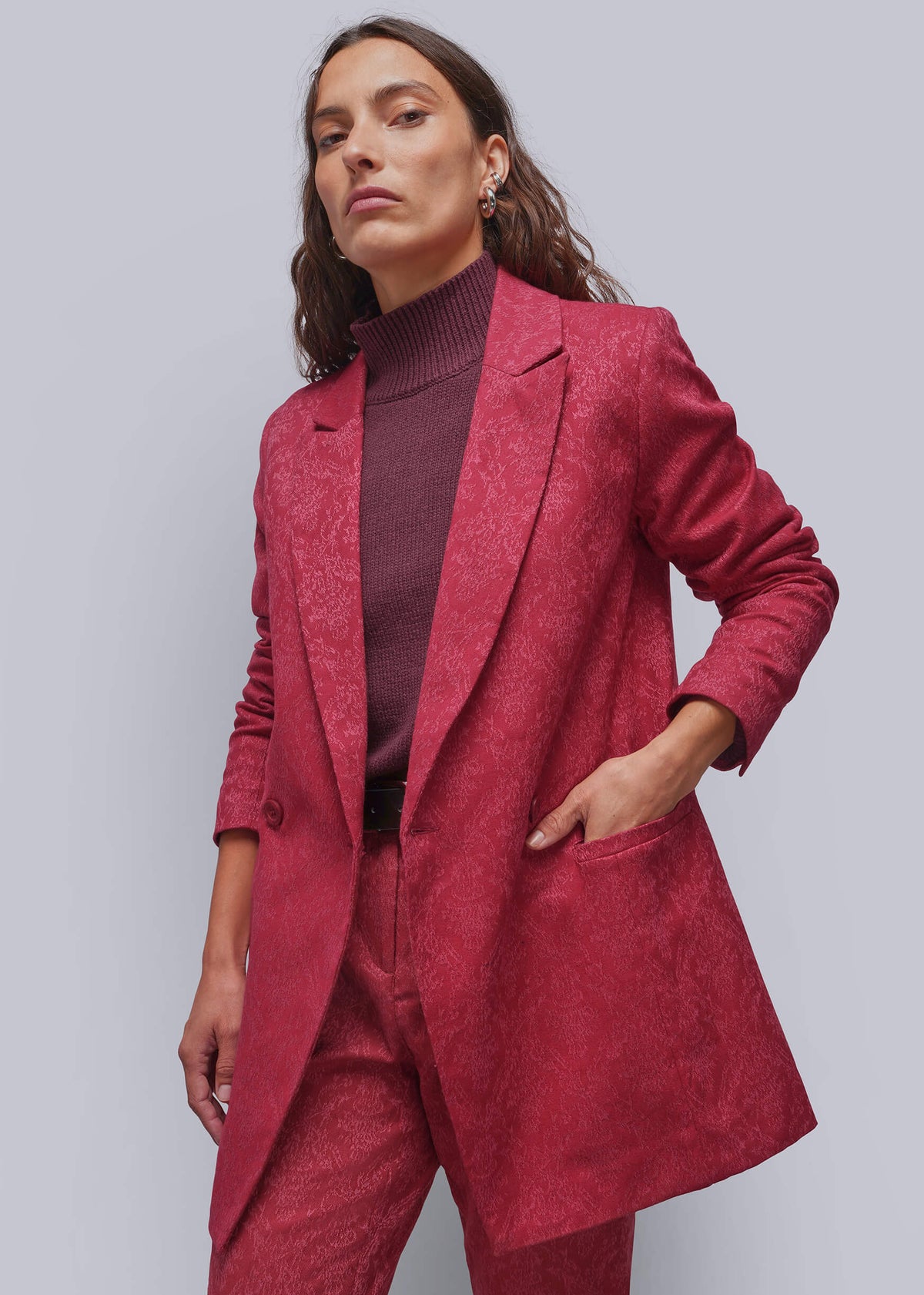 S=A woman modeling a matching textured red blazer and trousers set with a burgundy turtleneck sweater, photographed against a solid light gray studio background.