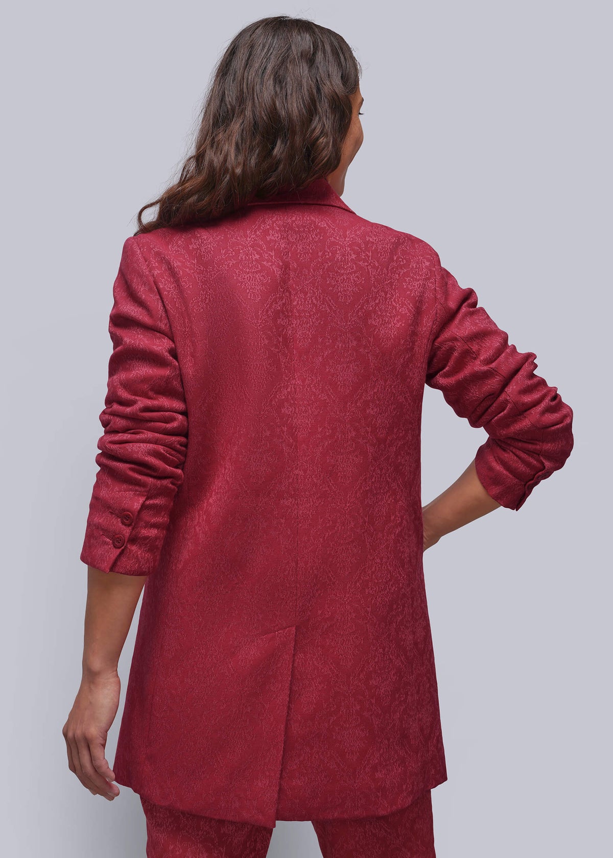 S=A woman shown from the back wearing a textured, deep red blazer and matching trousers against a solid gray studio background.