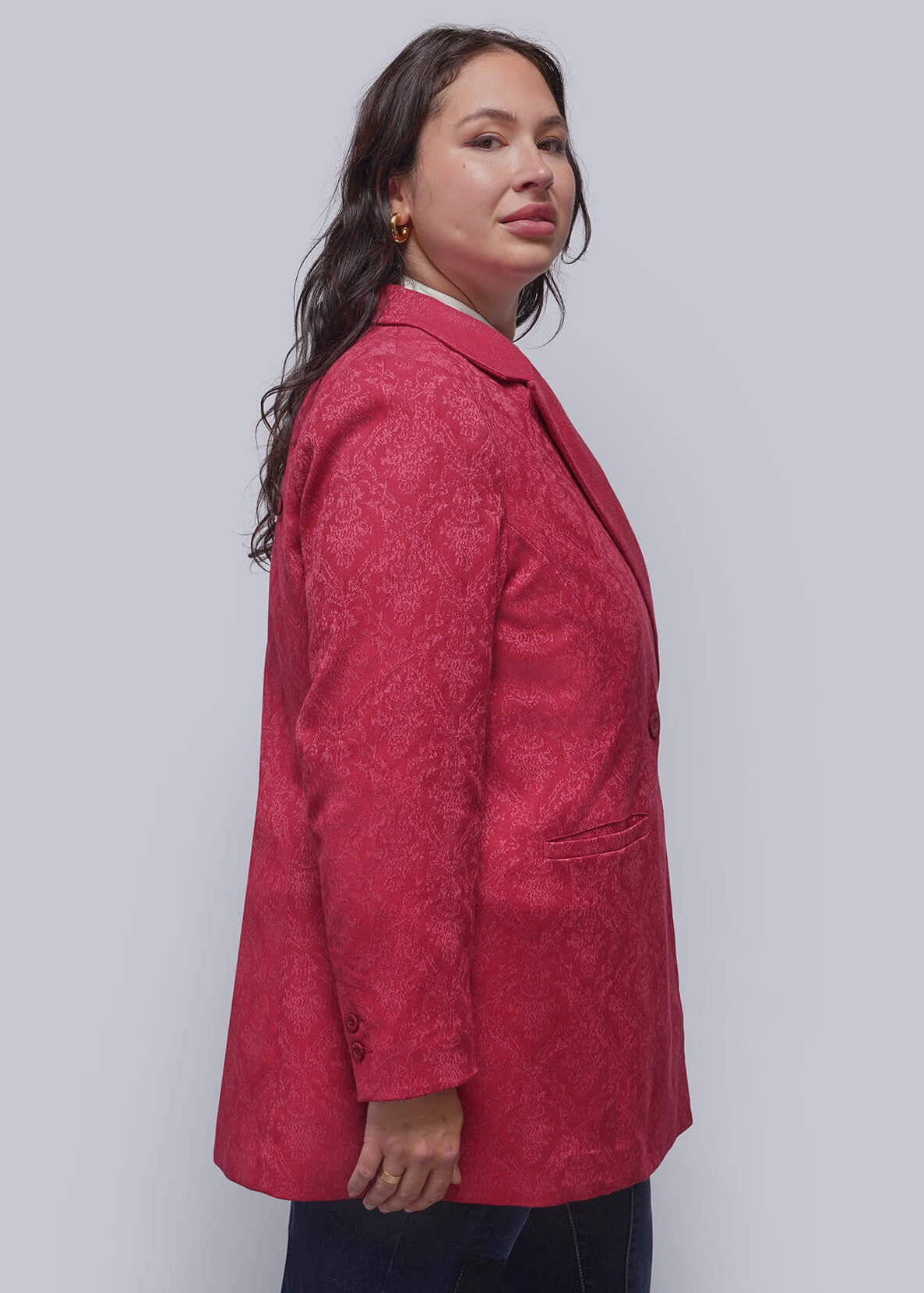 1X=A person modeling a red textured blazer, dark blue jeans, and black ankle boots, standing in profile against a light gray studio wall.