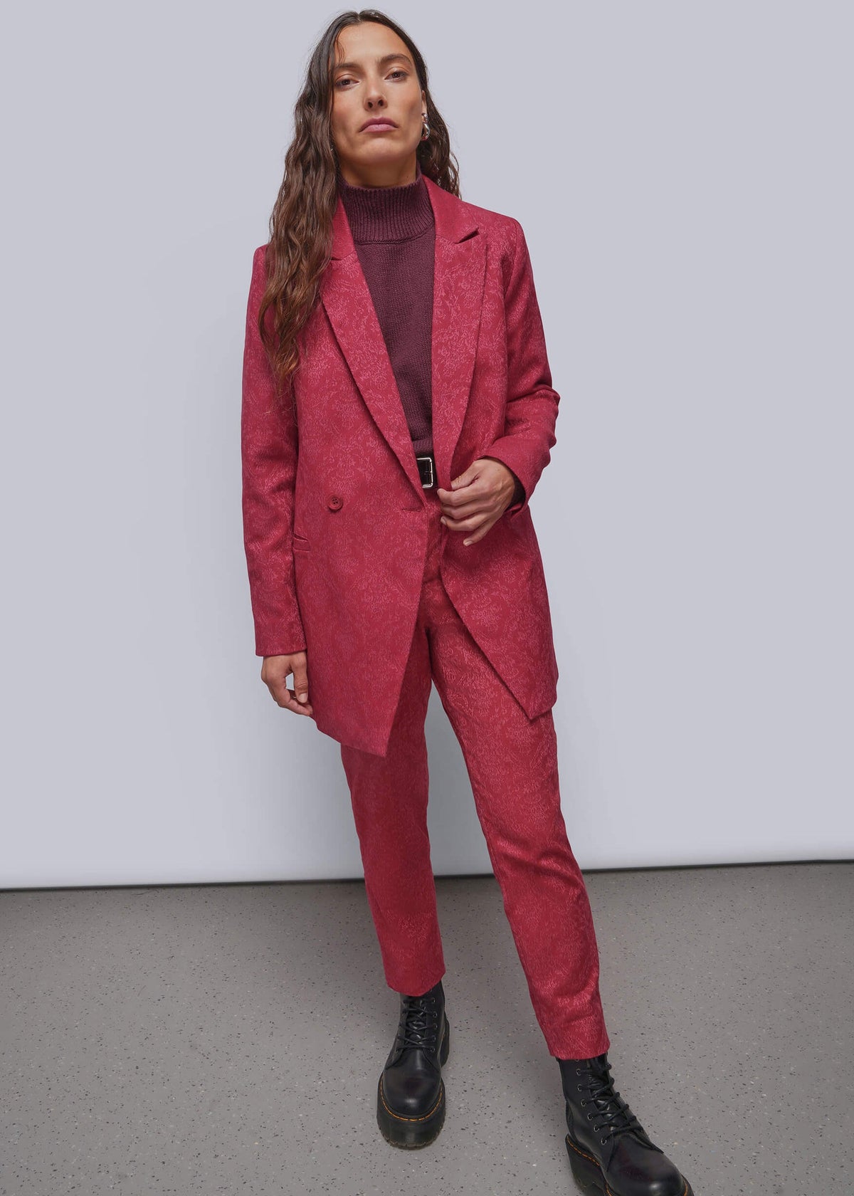 S=A woman modeling a matching textured red blazer and trousers suit with a dark burgundy turtleneck and black combat boots, standing against a light gray studio background.