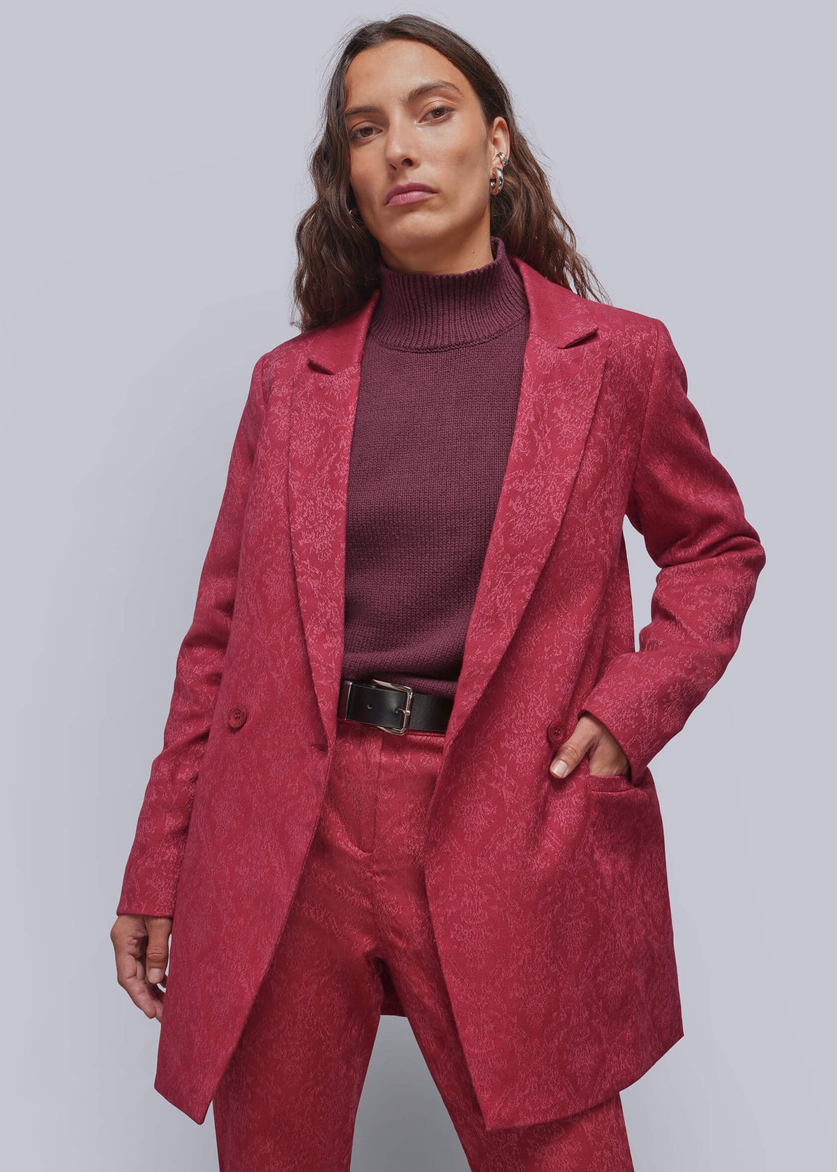 S=A model wearing a textured, jacquard pink blazer and matching trousers over a dark burgundy turtleneck sweater, styled with a black belt, against a solid gray studio background.