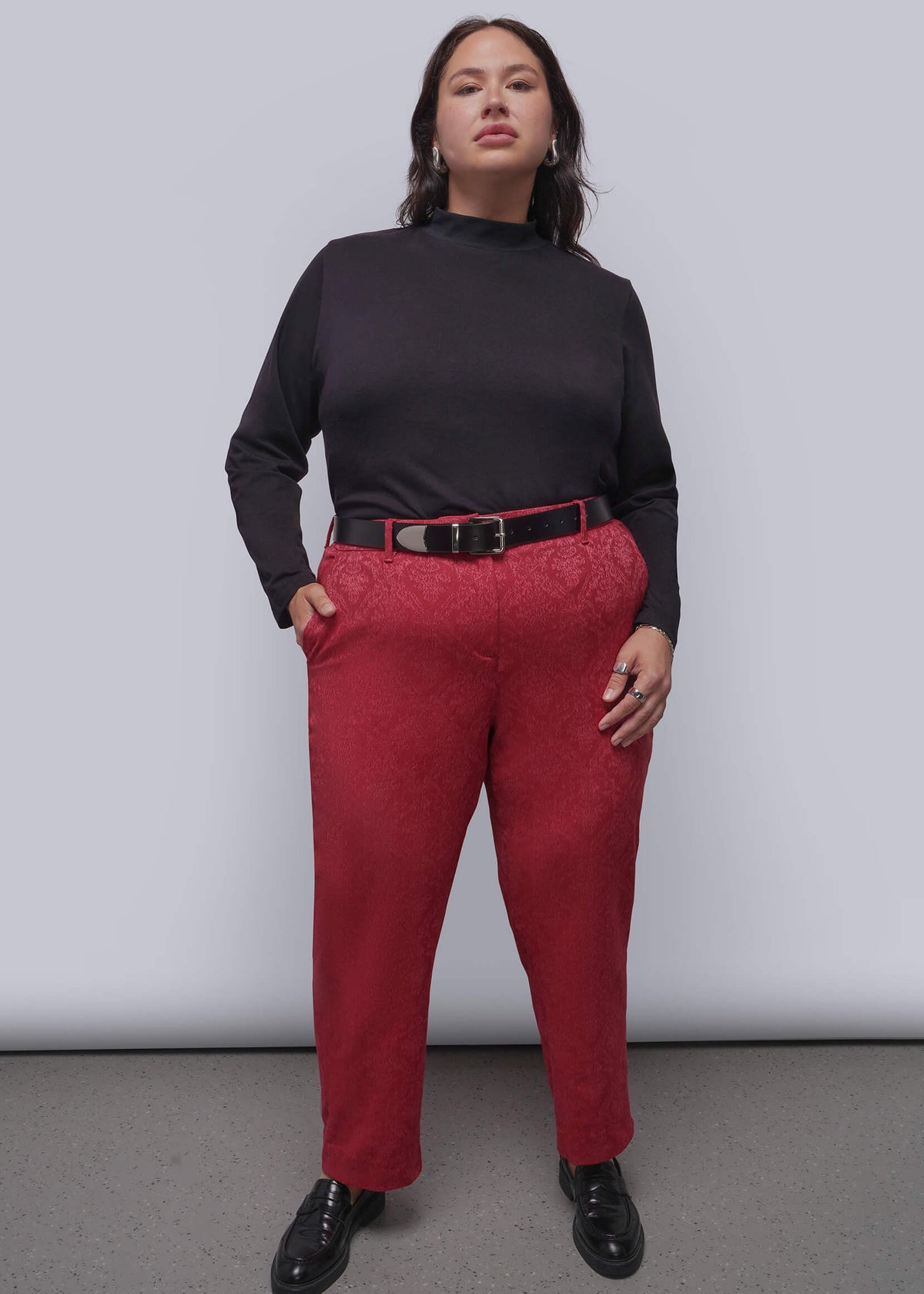 1X=A woman modeling a black mock-neck long-sleeve top and textured pink trousers with a black belt, standing against a light gray studio background.