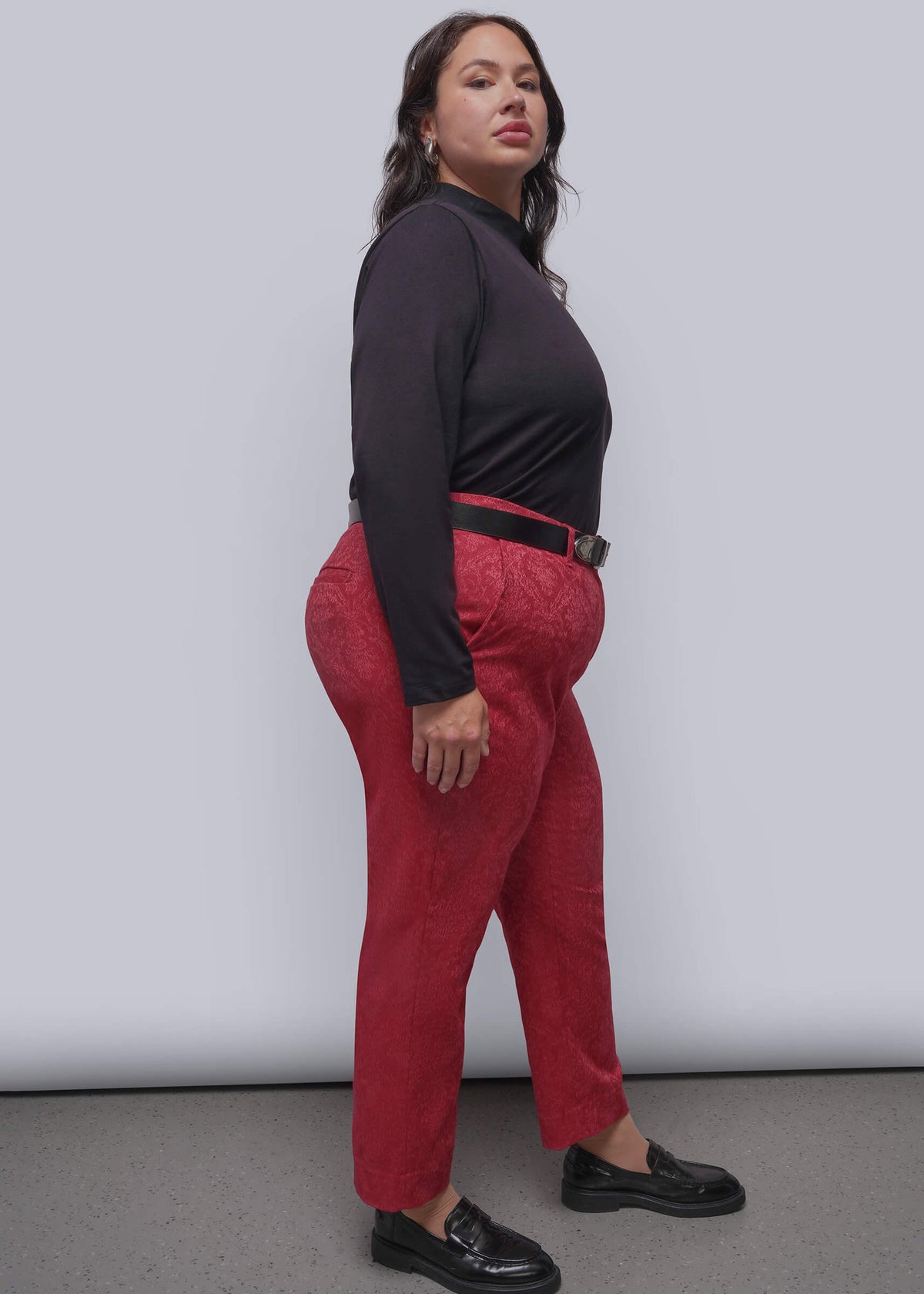 1X=A woman modeling bright pink textured trousers with a black belt, paired with a black long-sleeve top and black loafers, standing against a light gray studio background.