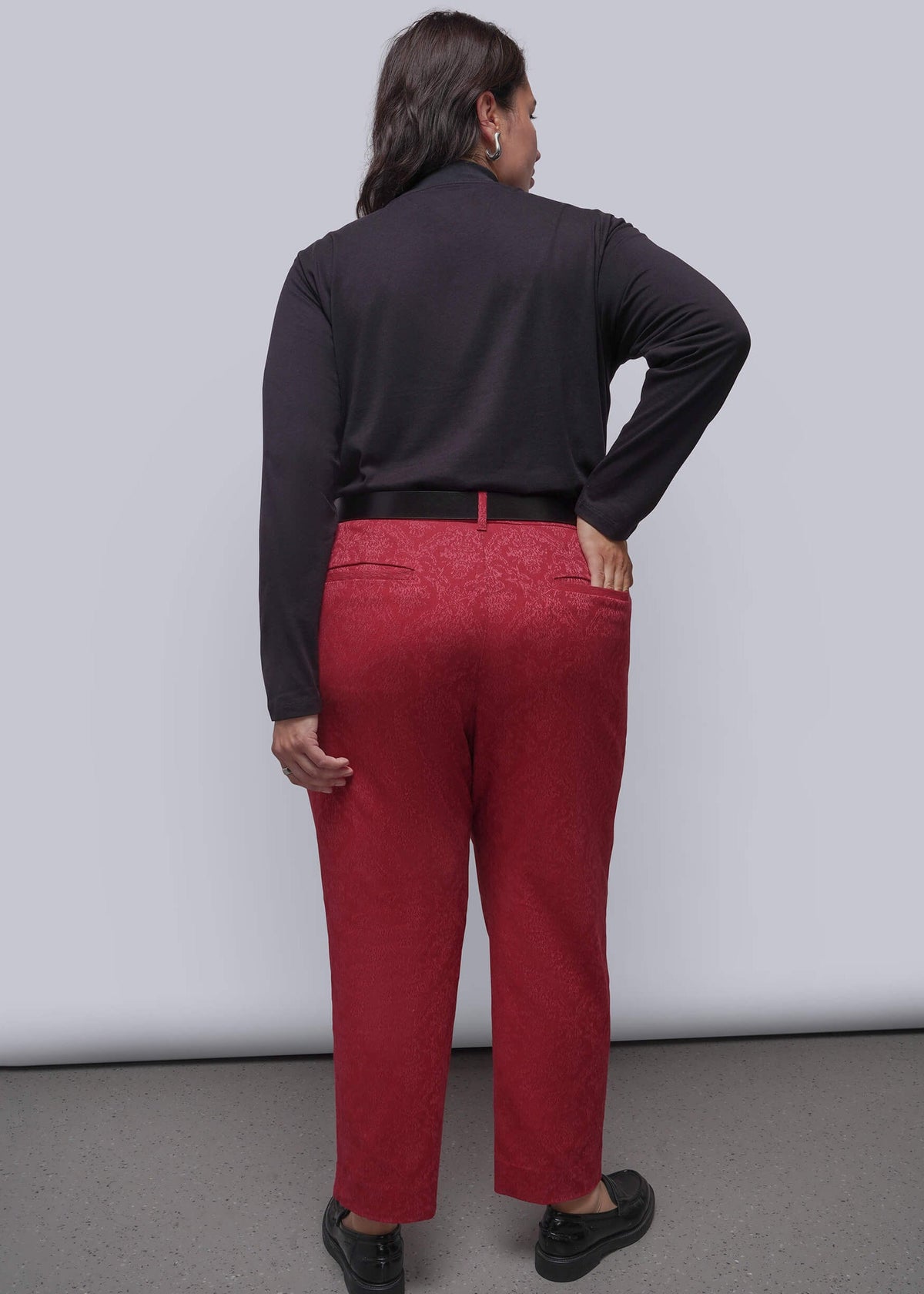 1X=A person shown from the back wearing bright pink textured trousers, a black long-sleeve top, and black shoes against a plain light gray studio background.