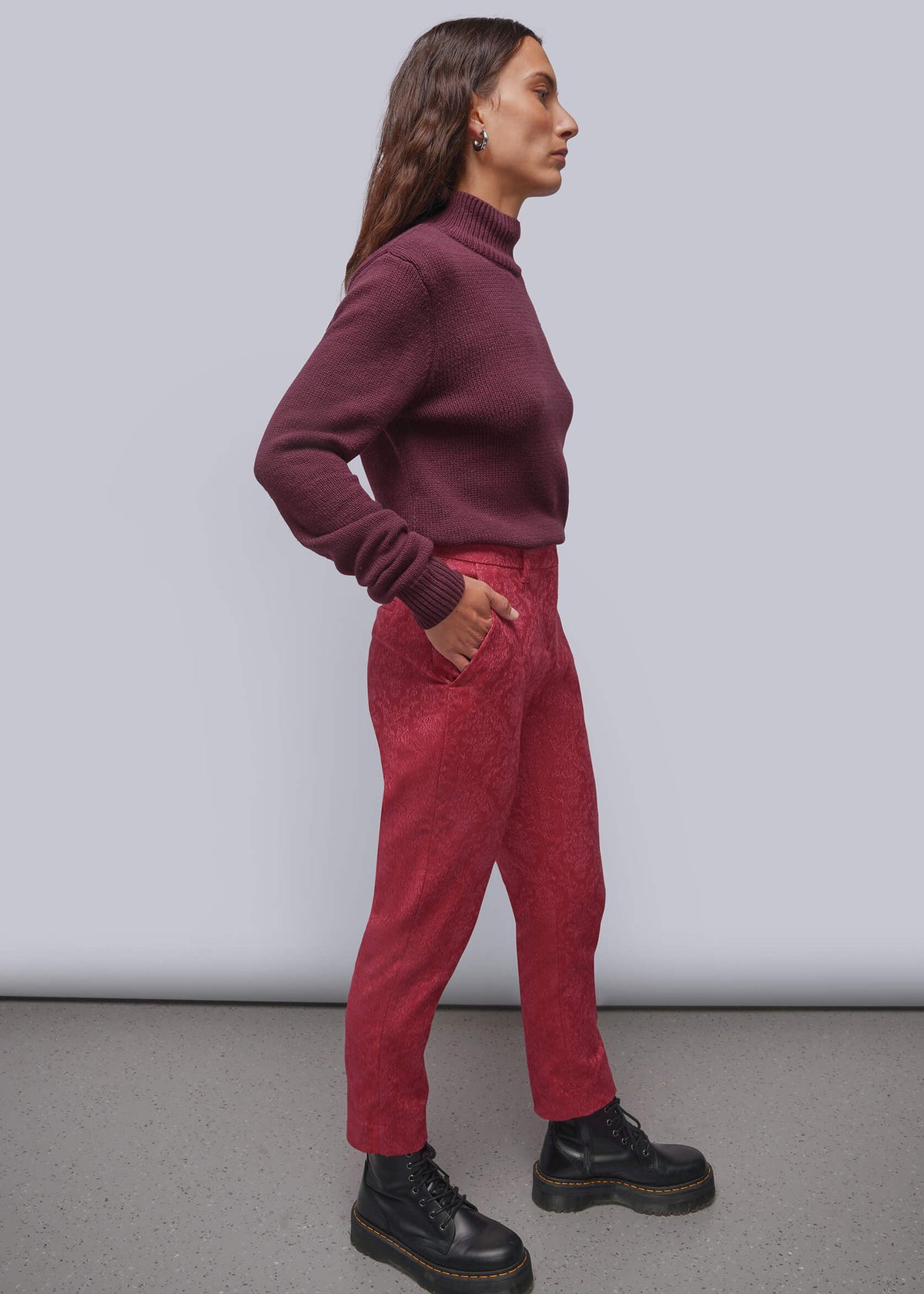 S=A woman stands in profile wearing a dark burgundy turtleneck sweater, textured pink pants, and black boots against a light gray studio background.