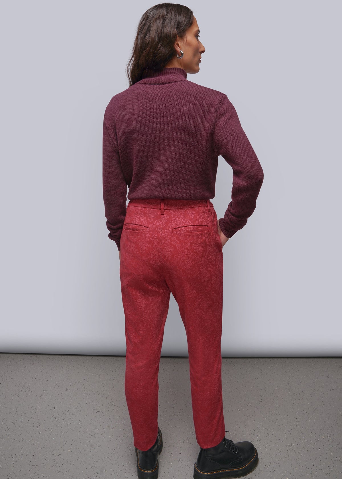 S=A woman is shown from behind wearing a dark burgundy mock-neck knit top and textured pink trousers, standing against a plain light gray studio wall.