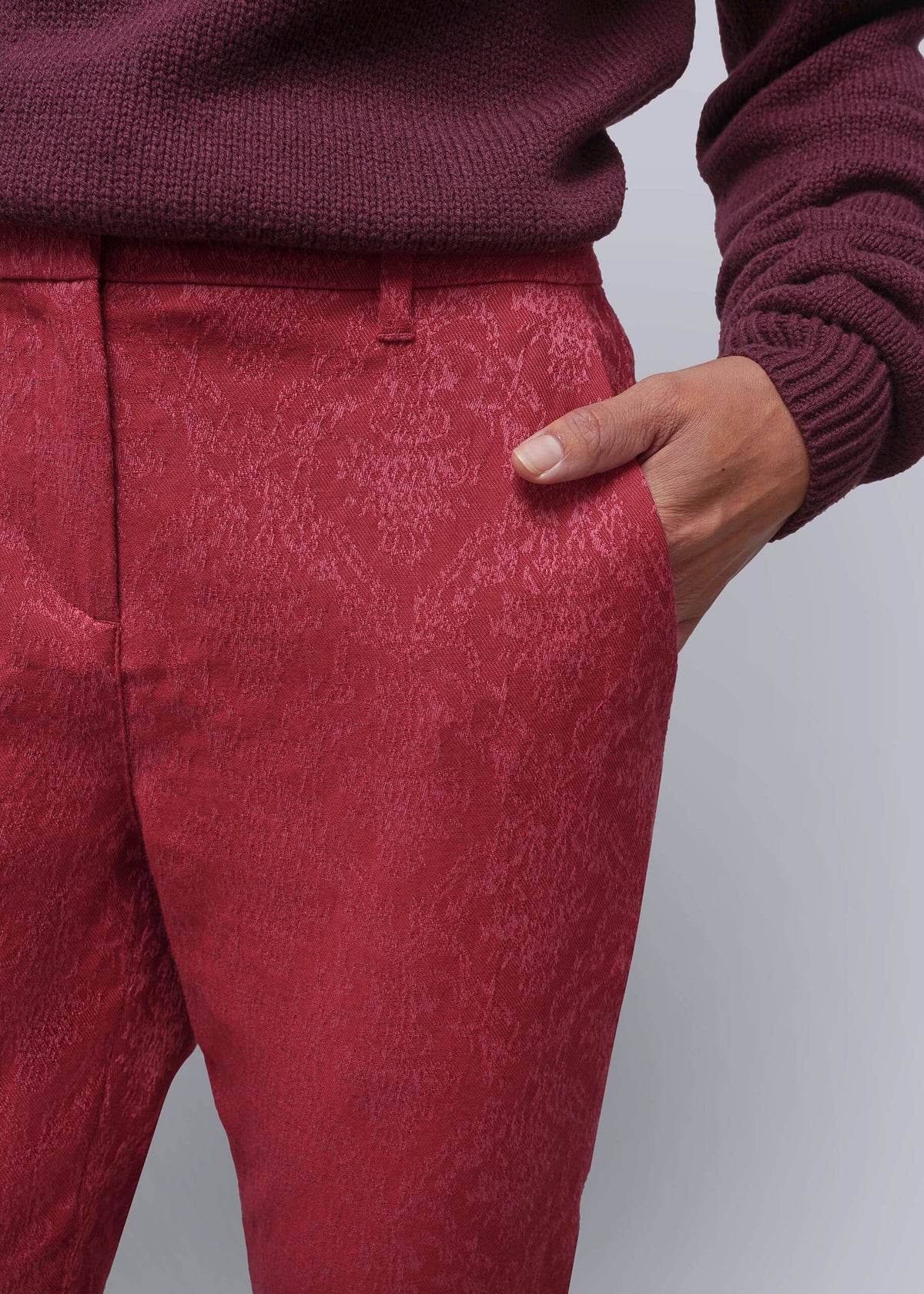 S=A close-up of a person wearing textured pink trousers and a matching burgundy knit sweater, with one hand in the pocket of the trousers, against a light gray background.
