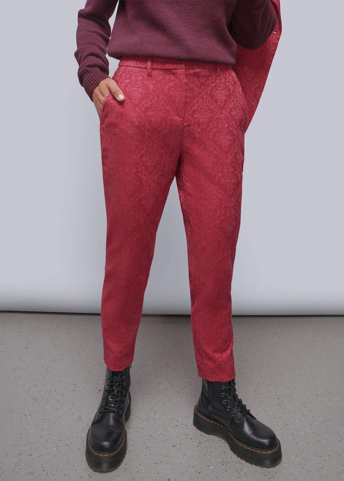 S=A person wearing textured red trousers, a burgundy sweater, and black leather boots, standing against a light gray studio background.