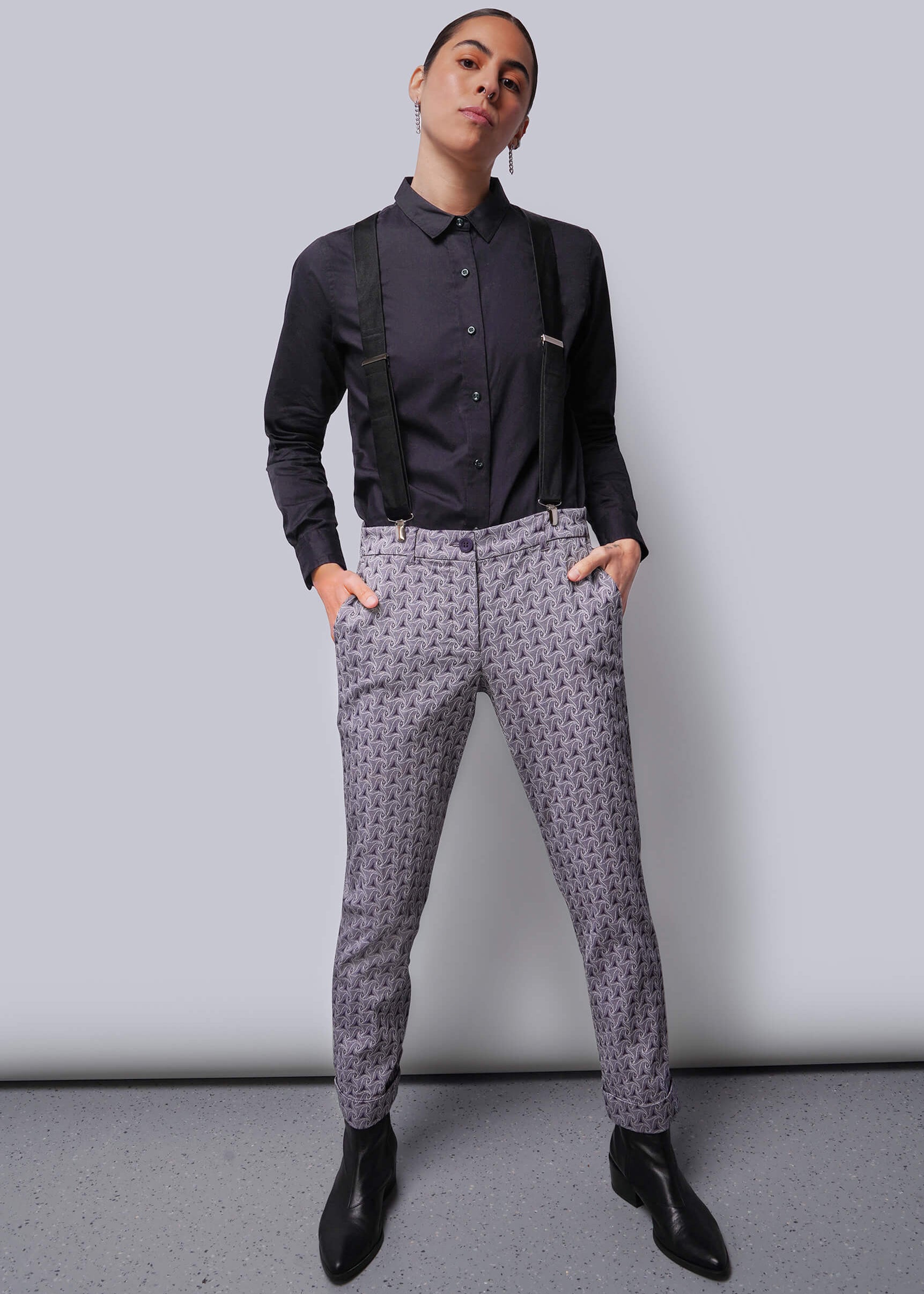 Person wearing a dark shirt, suspenders, and patterned pants against a plain background