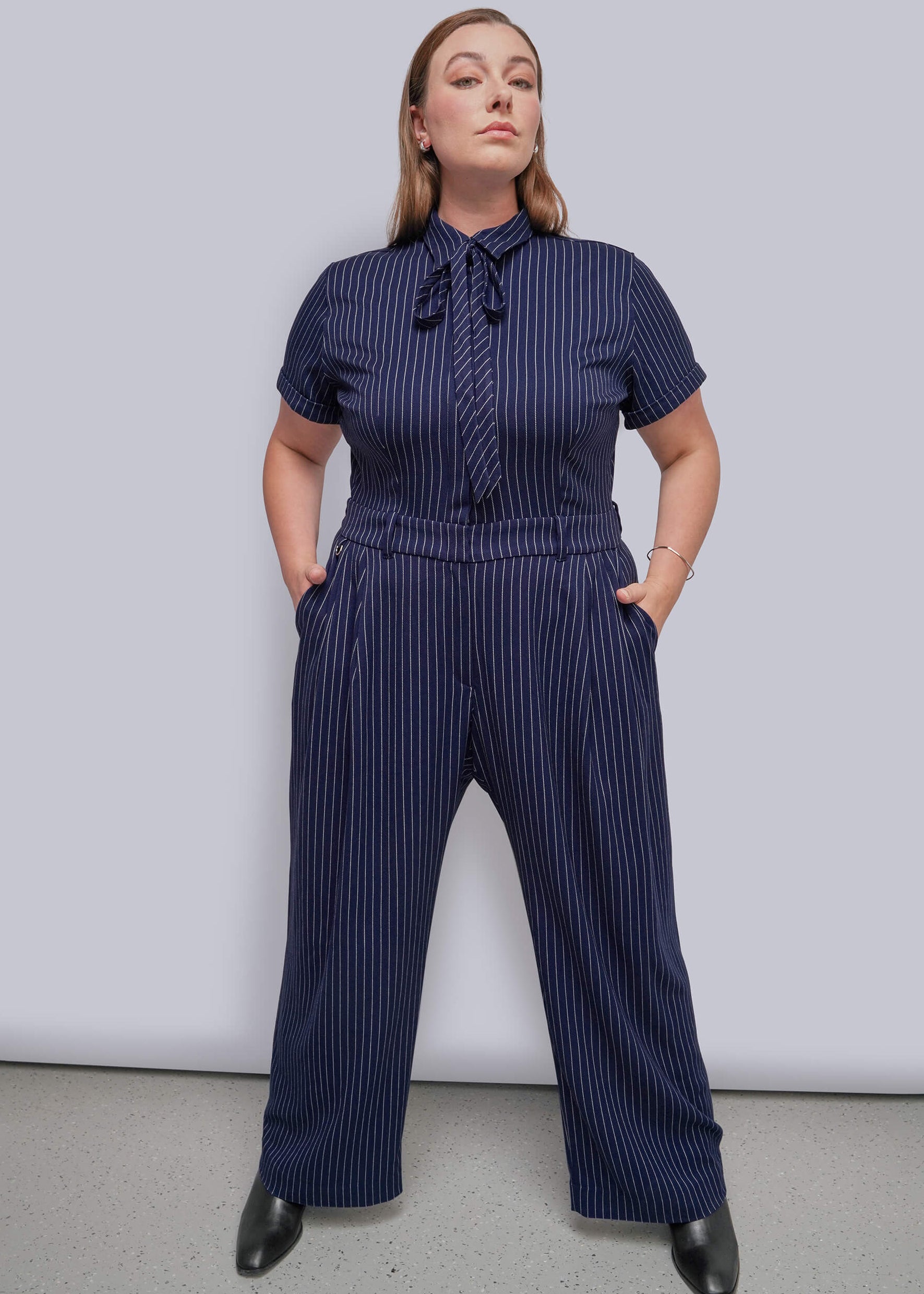 1X=A woman modeling a navy blue pinstriped short-sleeve jumpsuit with a tie-neck detail, standing against a light gray studio background.