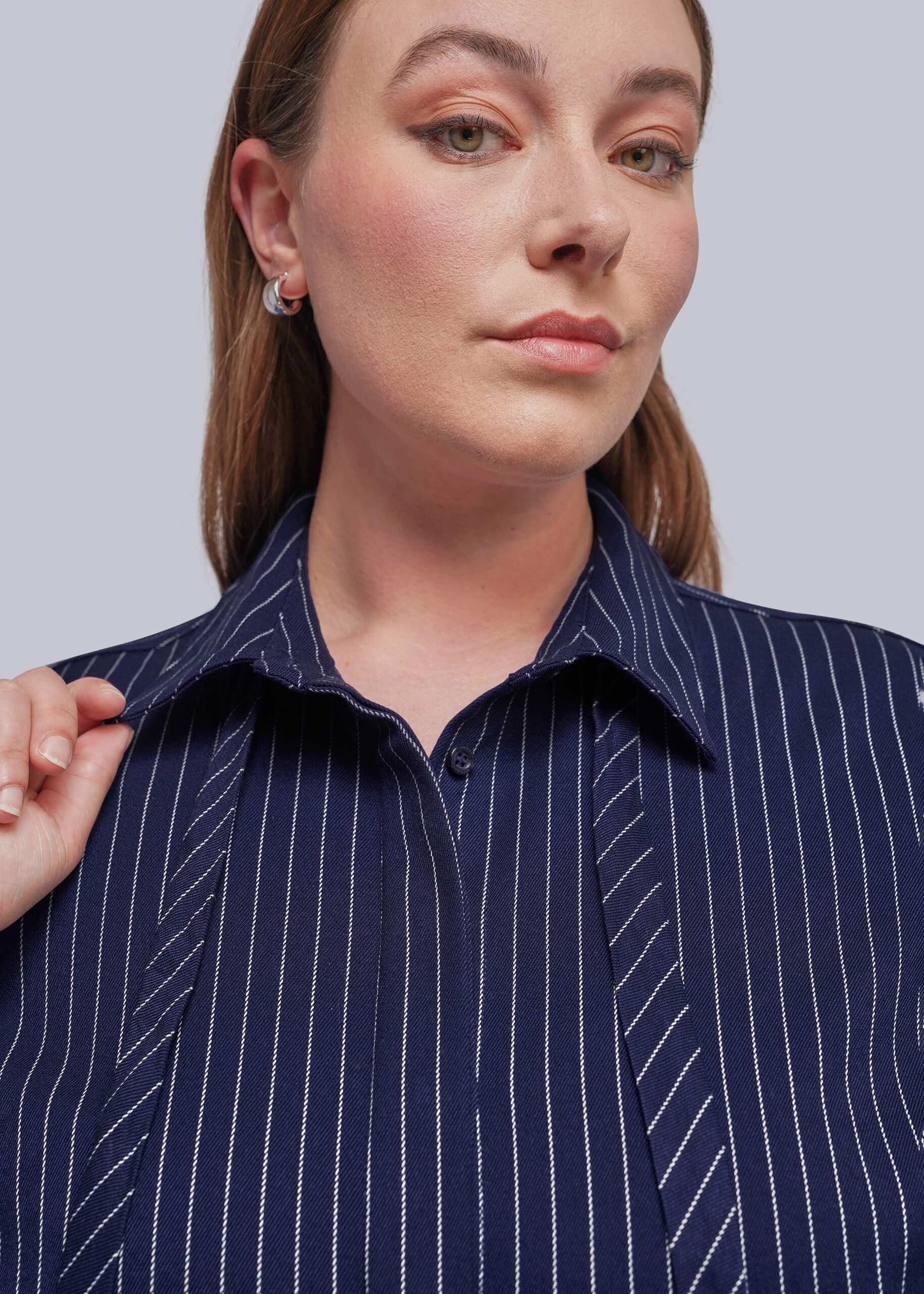 1X=A close-up of a woman wearing a navy blue button-up shirt with thin white vertical pinstripes, styled with a small silver hoop earring against a solid light gray background.