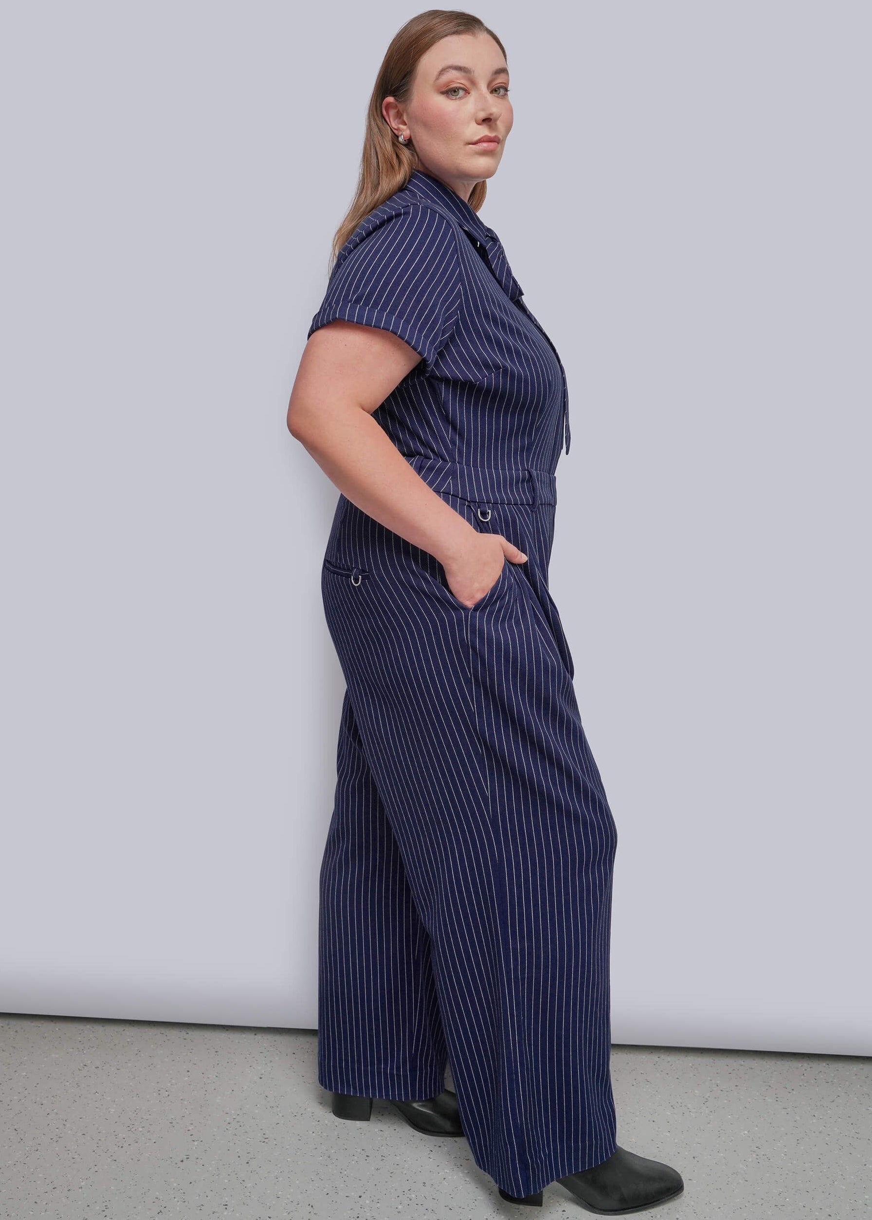 1X=A woman modeling a navy blue short-sleeve jumpsuit with thin white vertical pinstripes, standing against a light gray studio background.