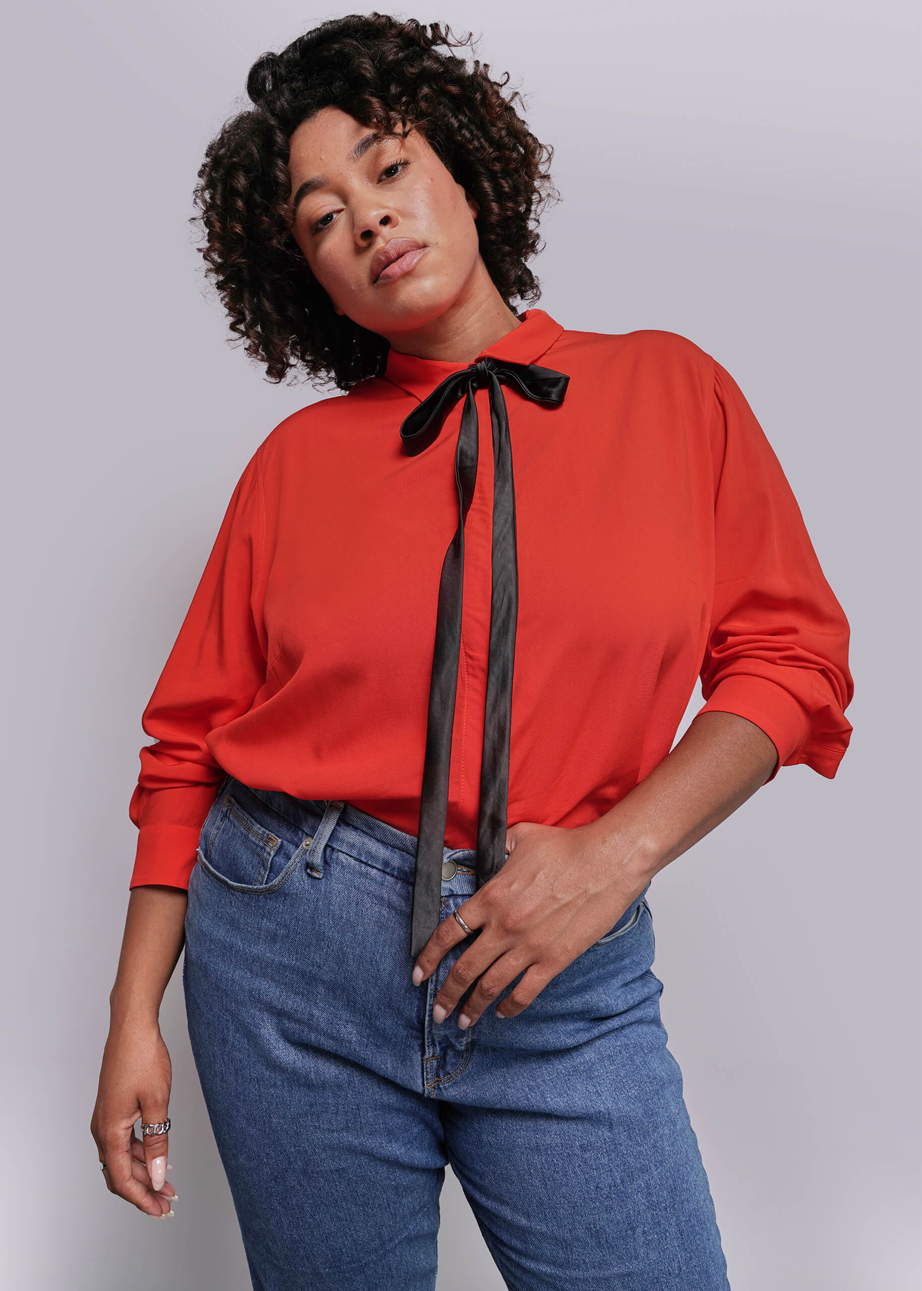 1X=Person wearing a red blouse with a black ribbon and blue jeans against a plain background