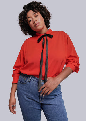 1X=Person wearing a red blouse with a black ribbon and blue jeans against a plain background