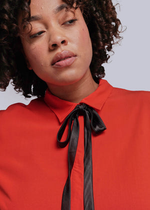 1X=Person wearing a red blouse with a black ribbon tie against a plain background