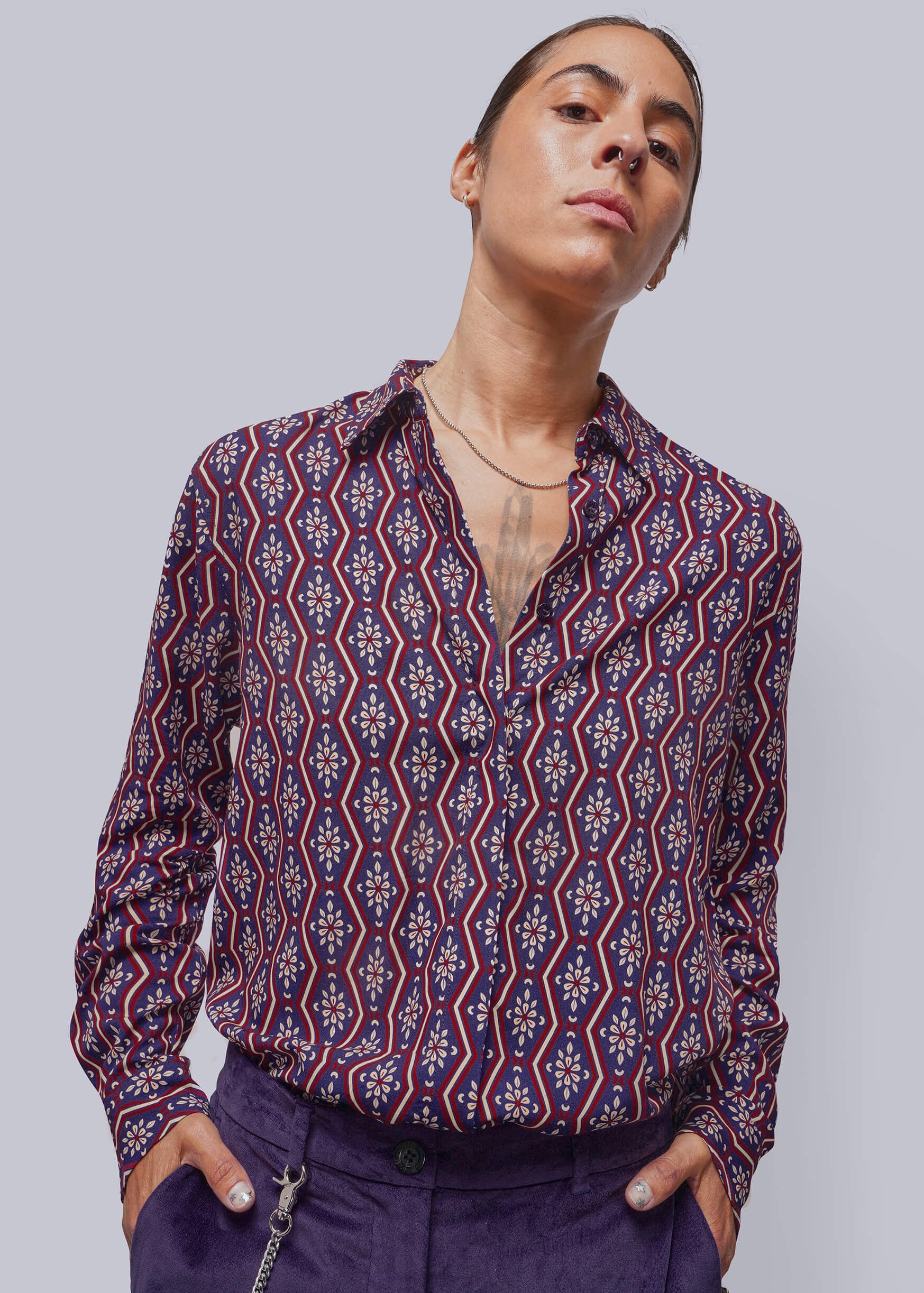A model wearing a long-sleeve button-up shirt with a red, blue, and white geometric pattern, paired with purple trousers, against a solid gray background.