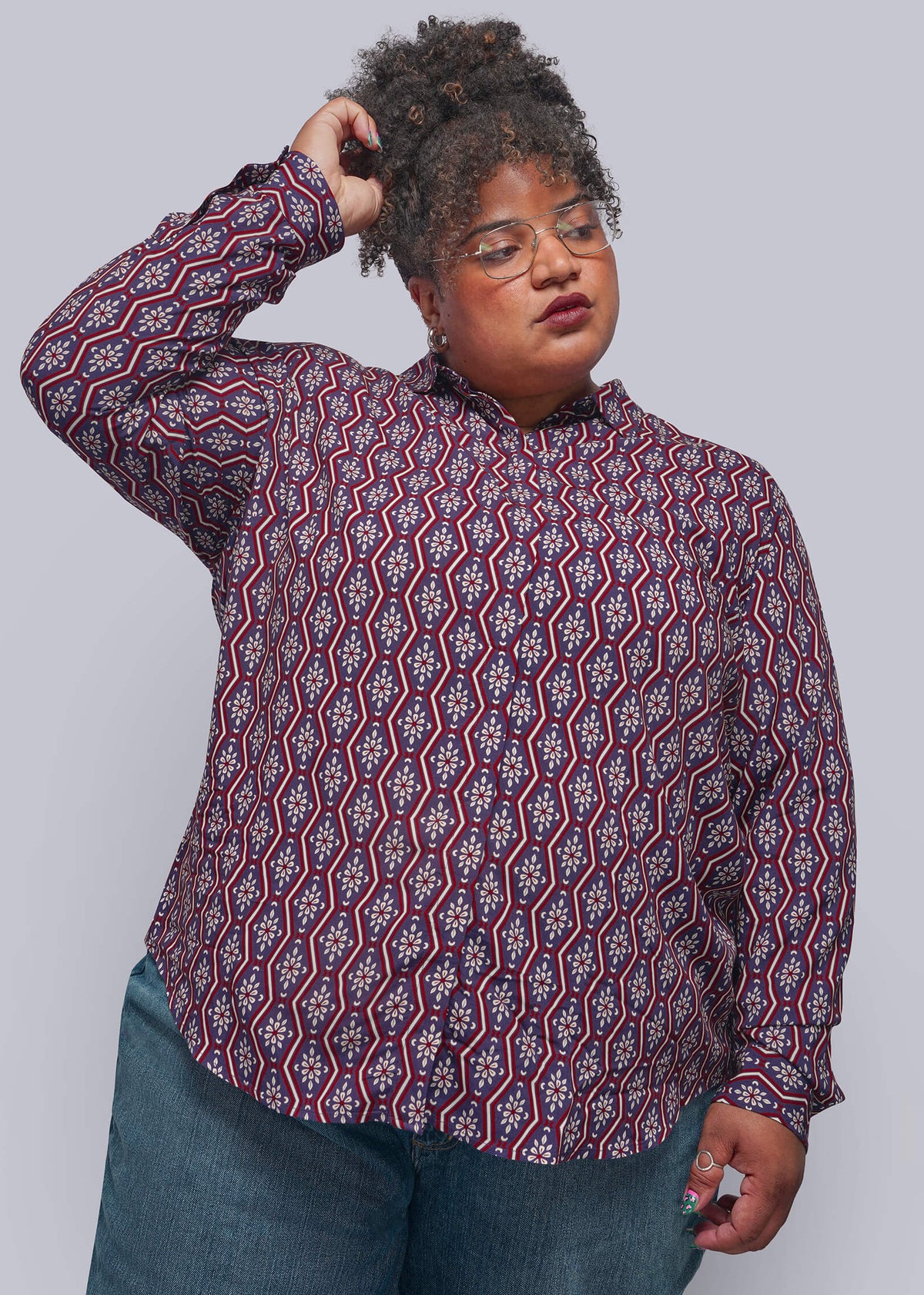 1X=A plus-size model wearing a long-sleeve button-up shirt with a red, purple, and white geometric pattern, paired with blue jeans, against a solid gray studio background.