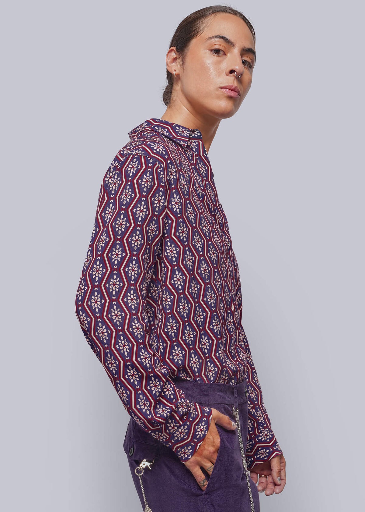 S=A model shown in profile wearing a long-sleeve patterned shirt with a purple, red, and white geometric design, paired with dark purple corduroy pants featuring a silver chain detail.