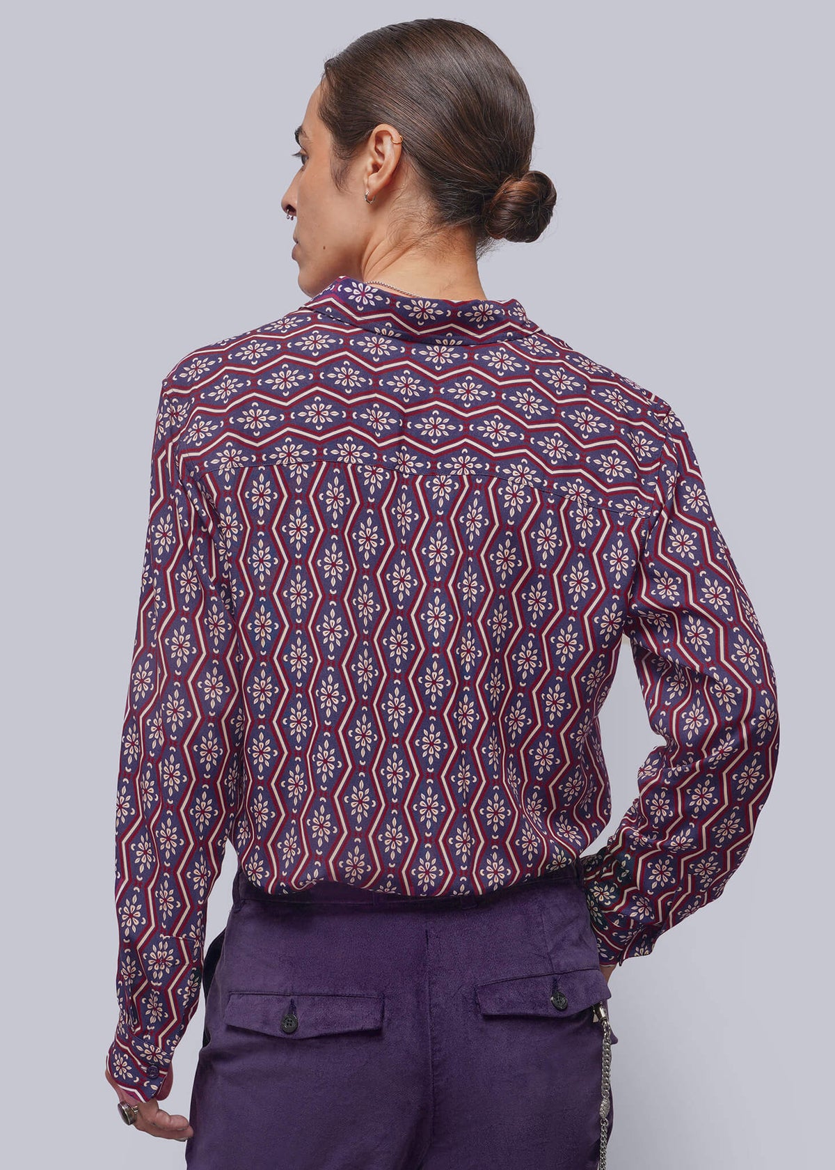 S=A person shown from the back wearing a long-sleeve button-up shirt with a red, purple, and blue geometric pattern, paired with solid purple trousers, against a solid gray studio background.