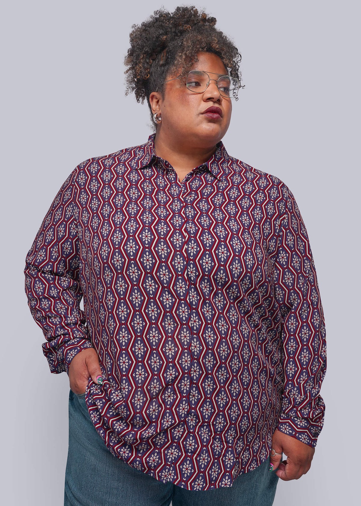 1X=A plus-size model wearing a long-sleeve button-down shirt with a red, blue, and white geometric pattern, paired with blue jeans, against a solid gray background.