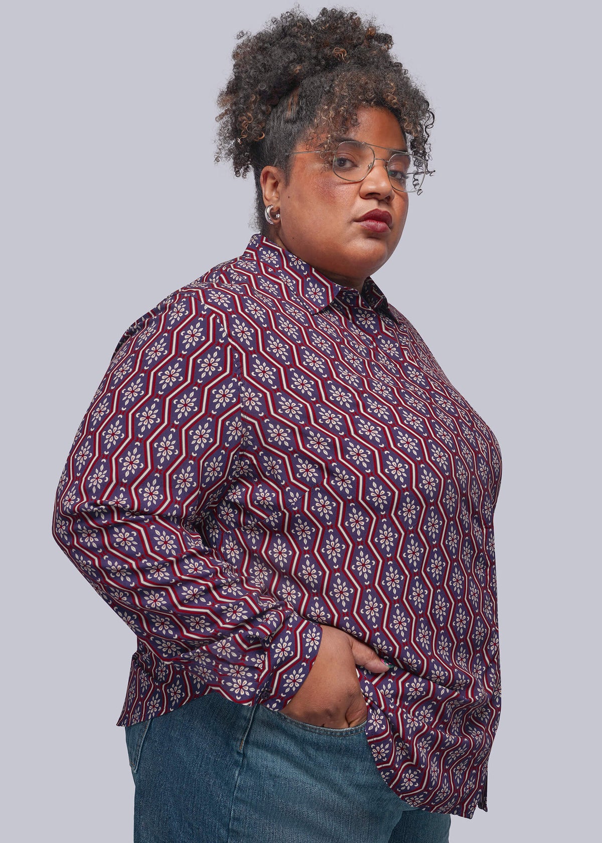 1X=A plus-size model wearing a long-sleeve button-down shirt with a red, navy, and white geometric pattern, paired with blue jeans, against a solid gray background.