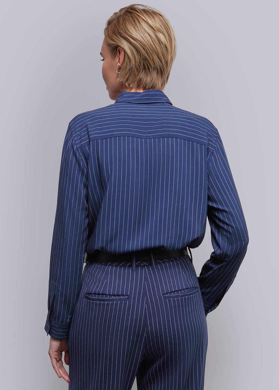 S=A model viewed from the back wearing a navy blue pinstriped shirt and matching trousers with a black belt, standing against a light gray studio background.