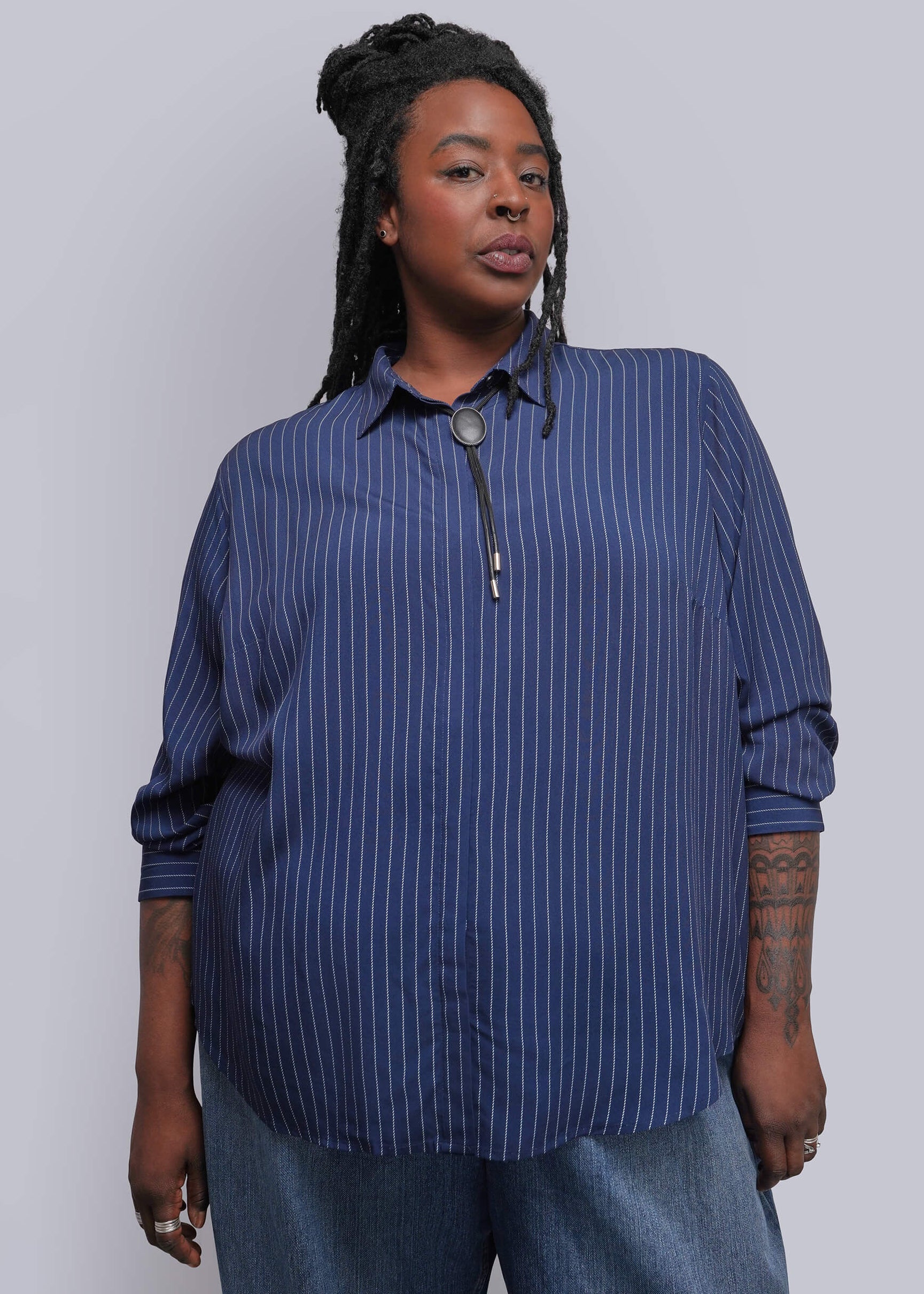 S=A woman modeling a blue and navy vertically striped button-down shirt with a front zipper, paired with blue jeans, against a plain light gray studio background.