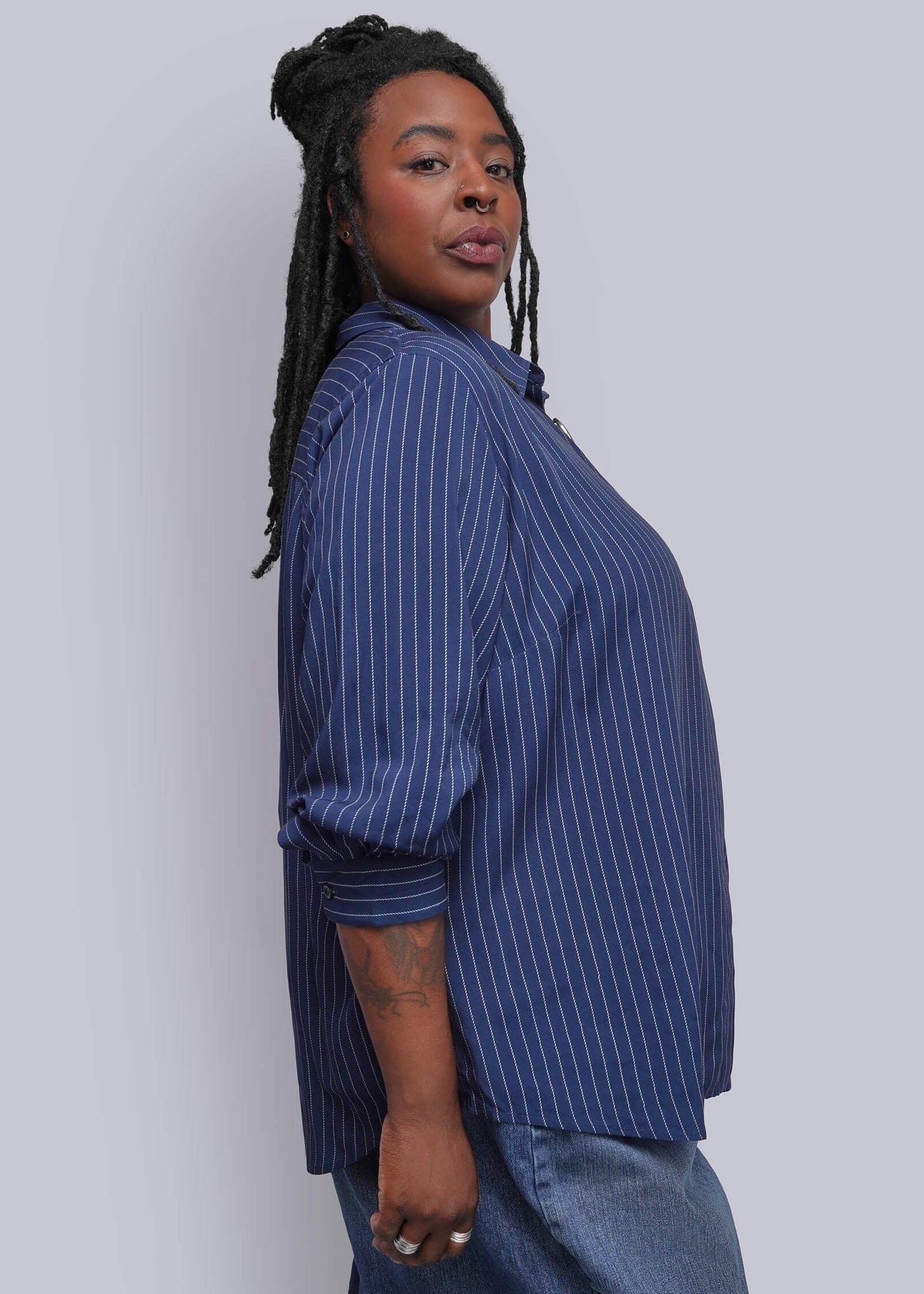 1X=A person with long black braids is shown from the side, wearing a blue and white vertically striped button-up shirt and blue denim pants against a plain light gray background.