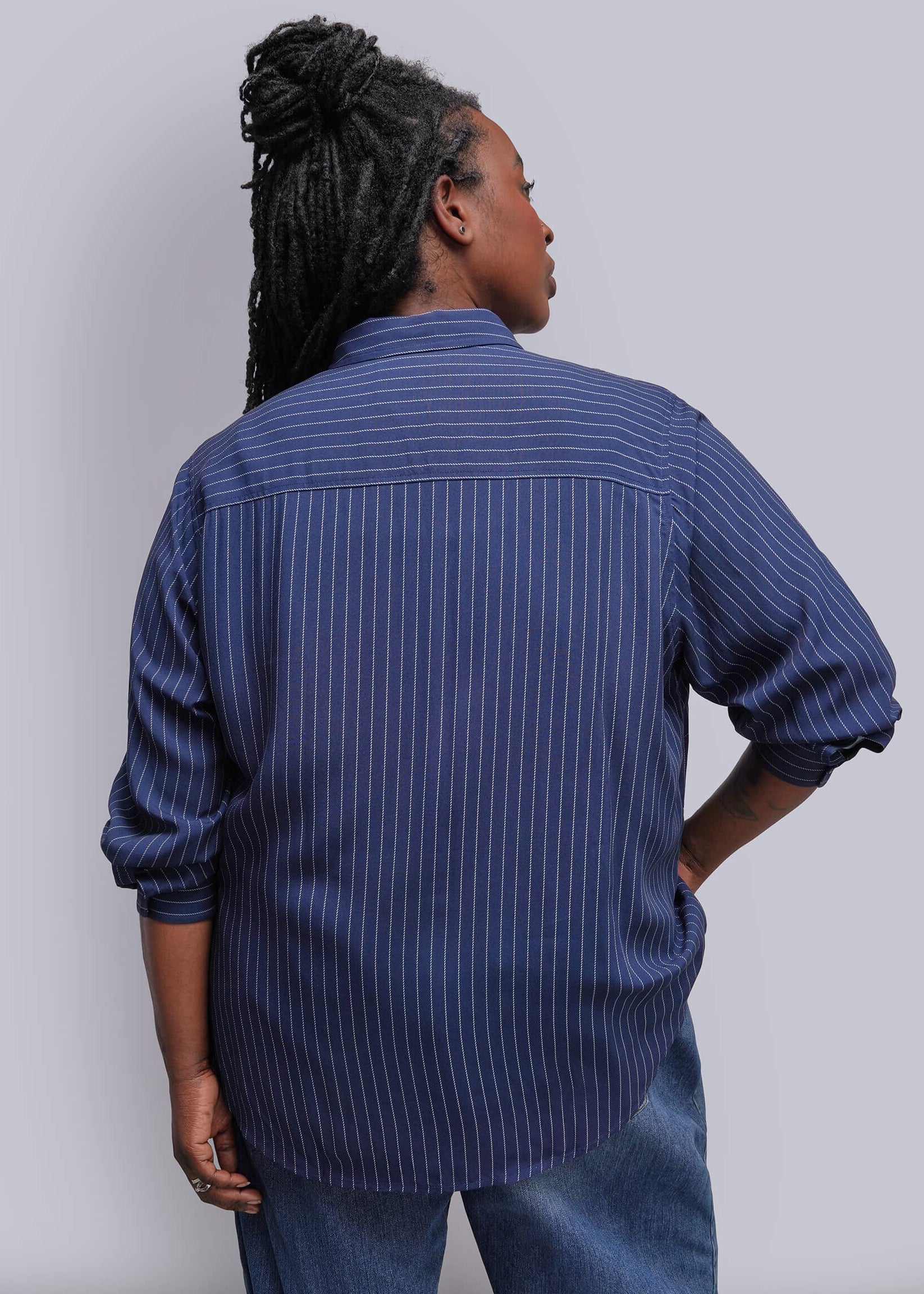 1X=A person shown from the back wearing a blue button-up shirt with thin white vertical pinstripes and blue jeans, standing against a plain light gray studio background.