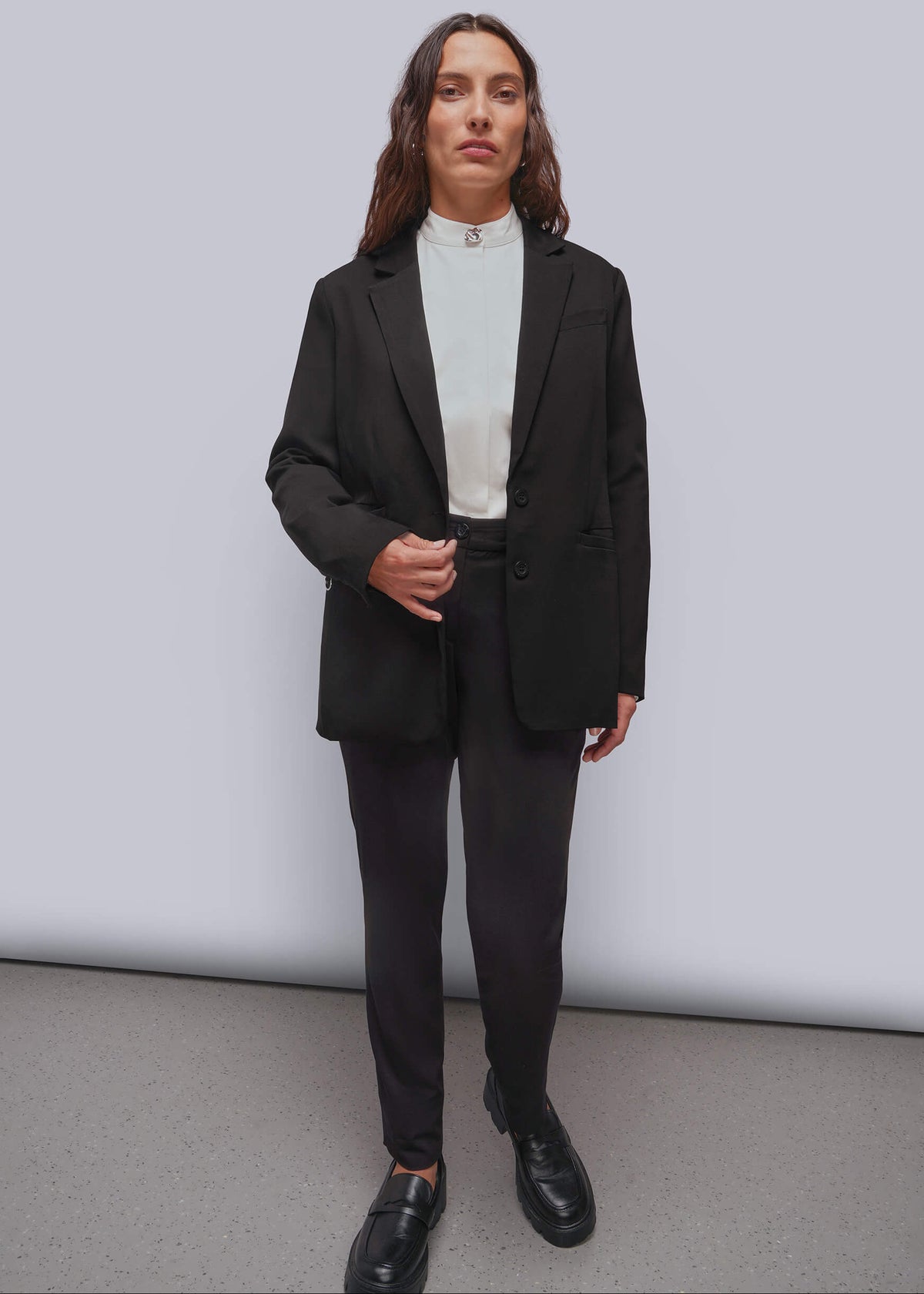 S=A model stands against a light gray studio wall wearing a black blazer, black trousers, a white button up, and black loafers.