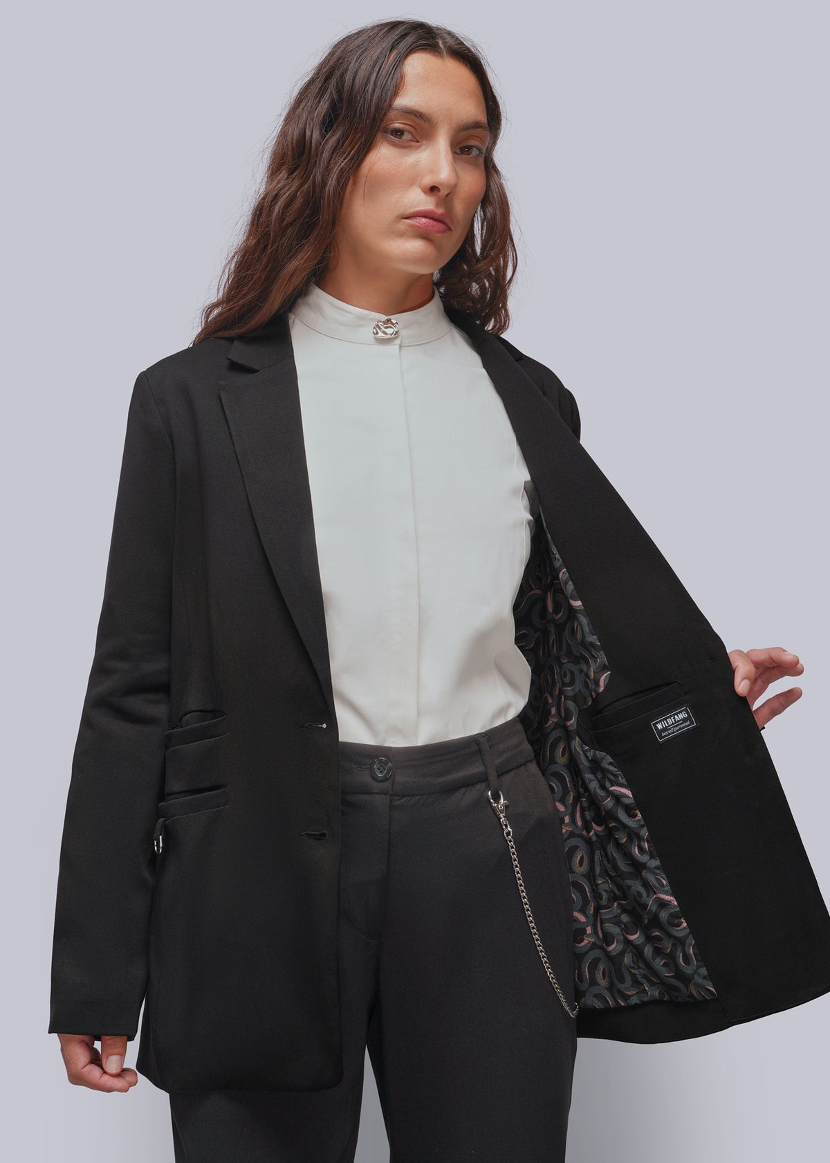 S=A model wearing a black blazer over a white button up top and dark trousers, holding open the blazer to show a patterned lining. The blazer has a small black brand patch on the pocket.