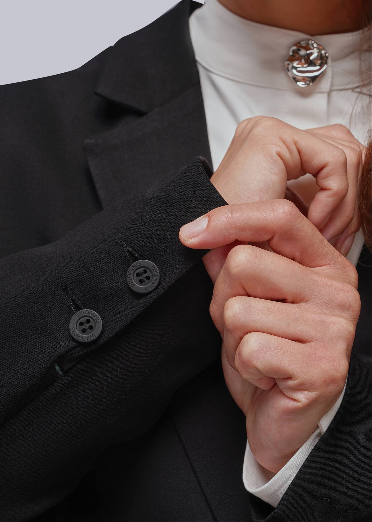 S=Close-up of a person wearing a black tailored blazer over a white high-neck top, with one hand adjusting the cuff and the other resting near the chest. A silver decorative button cover is visible at the neckline.