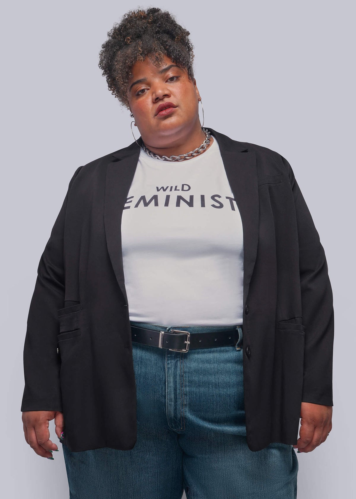 1X=A plus-size model wearing a black blazer over a white graphic t-shirt that reads 'WILD FEMINIST', paired with blue jeans and a black belt, against a solid gray studio background.