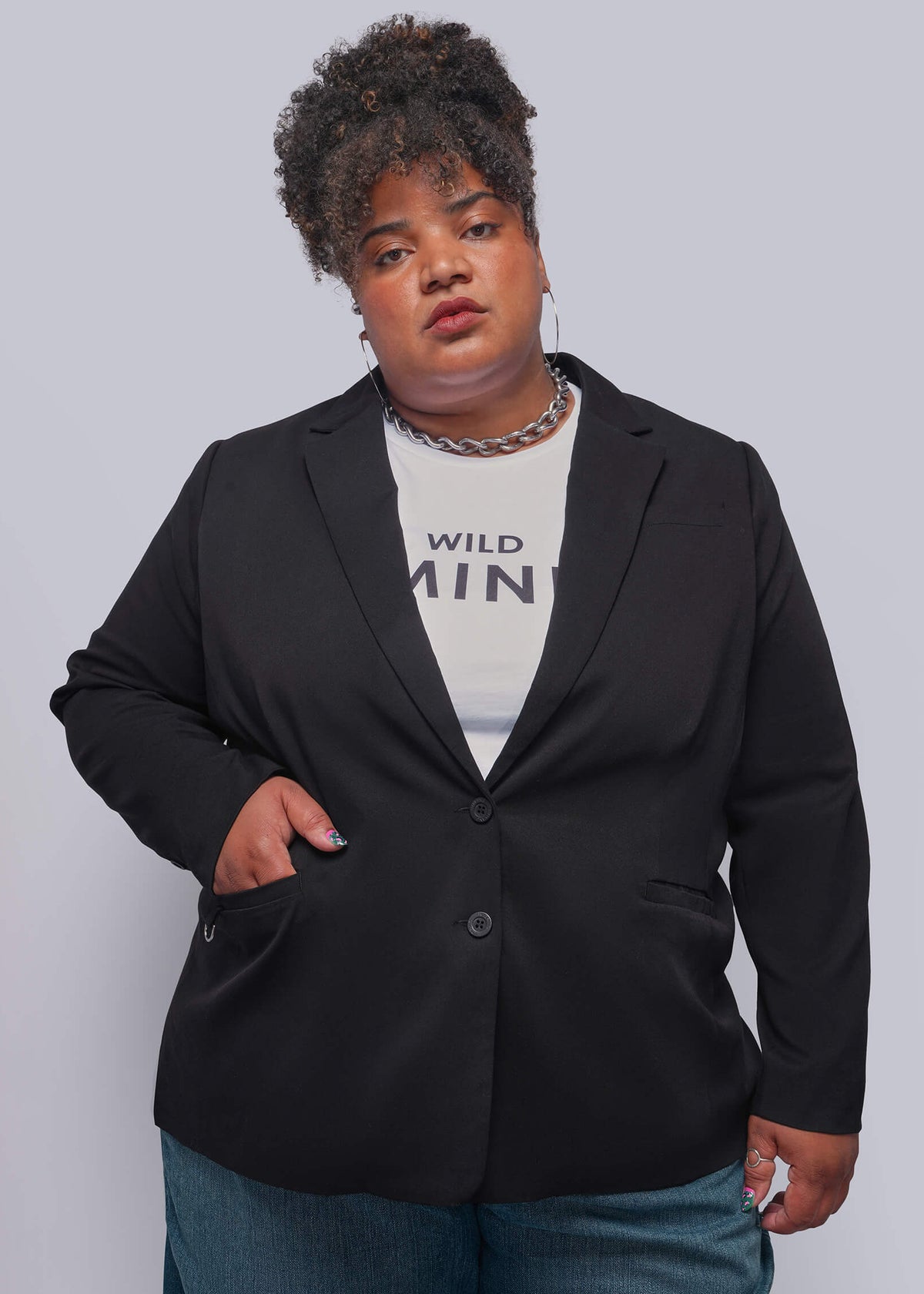 1X=A plus-size model wearing a black blazer over a white graphic t-shirt and blue jeans, standing against a solid gray studio background.
