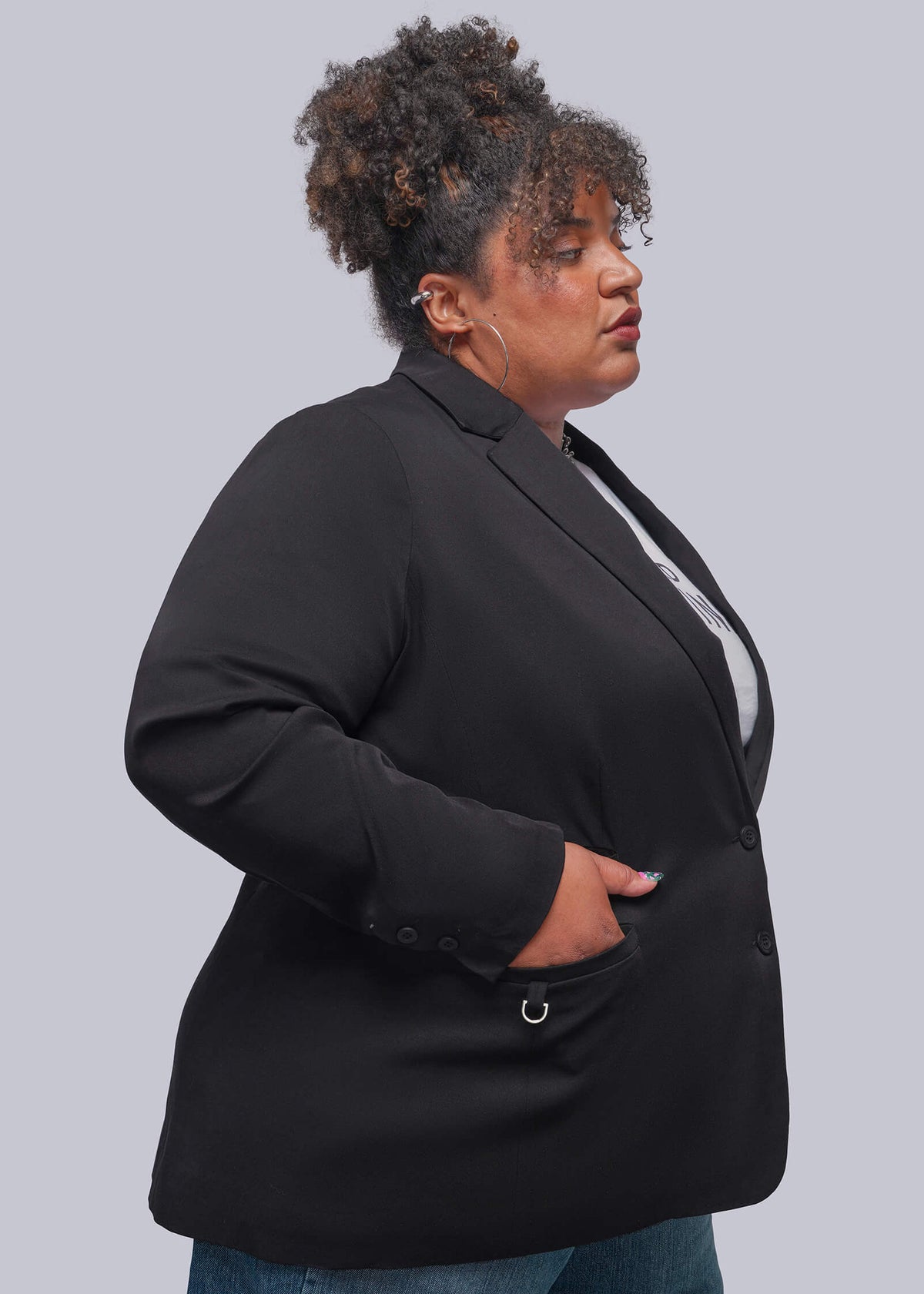 1X=A plus-size model wearing a black blazer with a silver ring detail on the pocket, paired with blue jeans, shown from a side profile against a solid gray background.