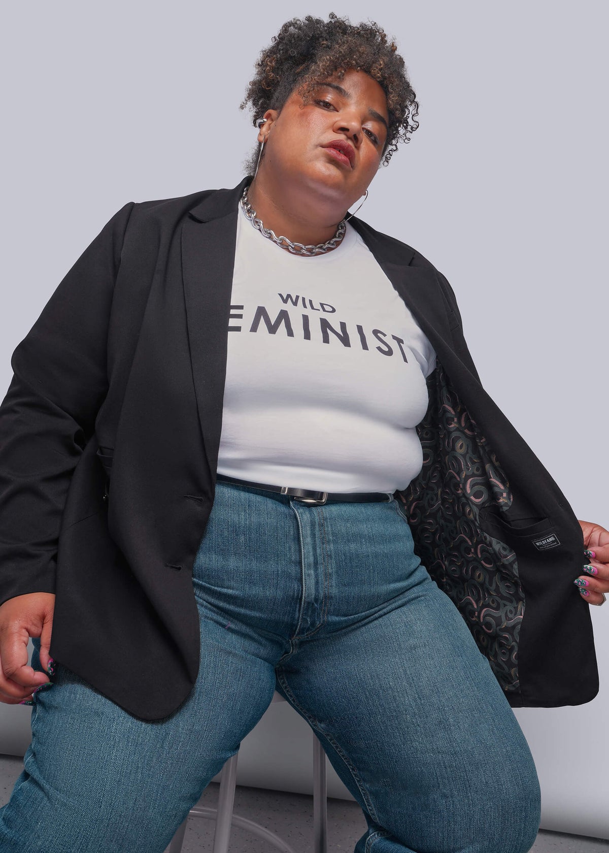 1X=A plus-size model wearing a black blazer over a white graphic t-shirt that reads 'WILD FEMINIST', paired with blue jeans, posing against a light gray studio background.