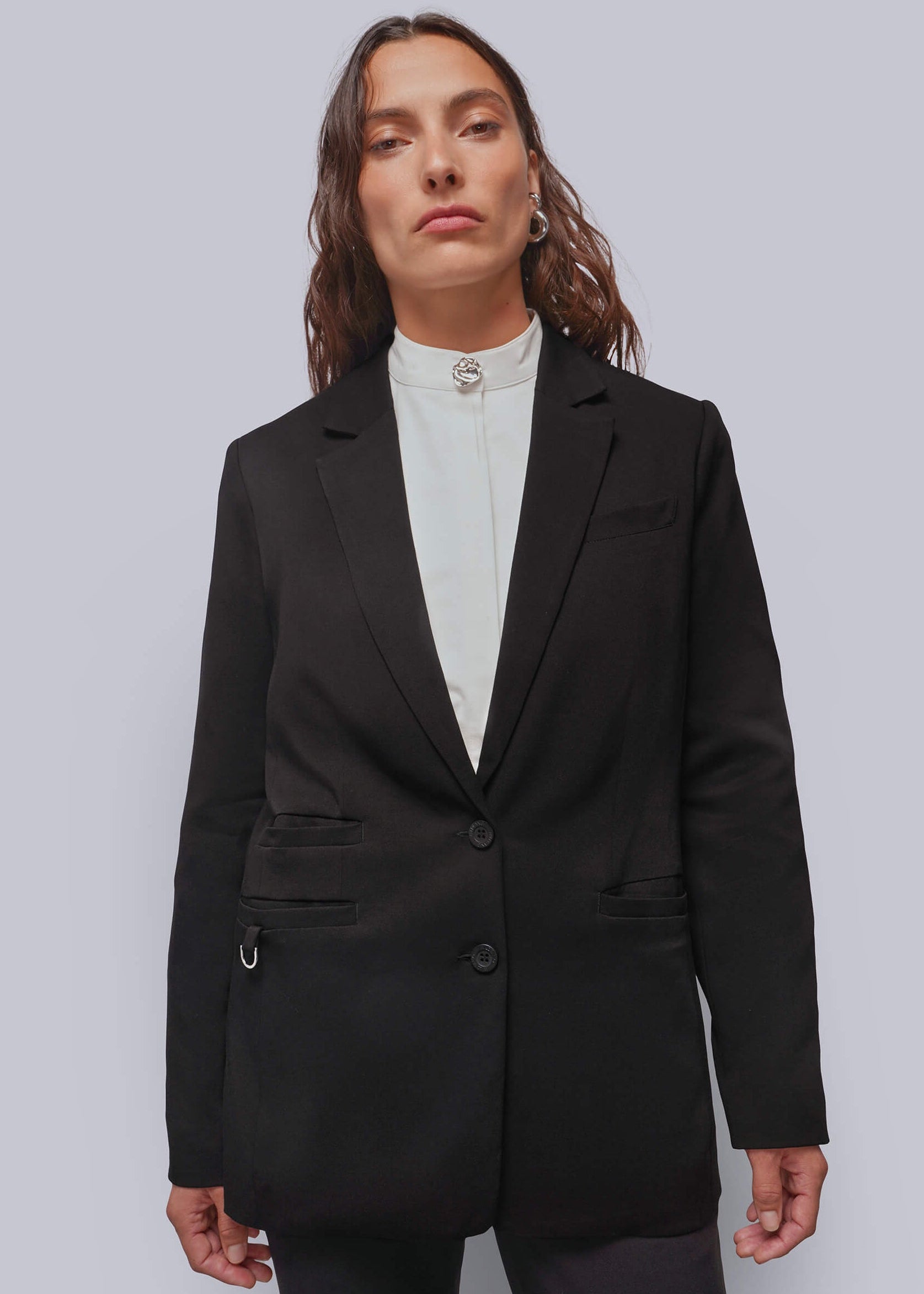 A woman wearing a black tailored blazer over a white mock-neck top, photographed against a plain light gray studio background.