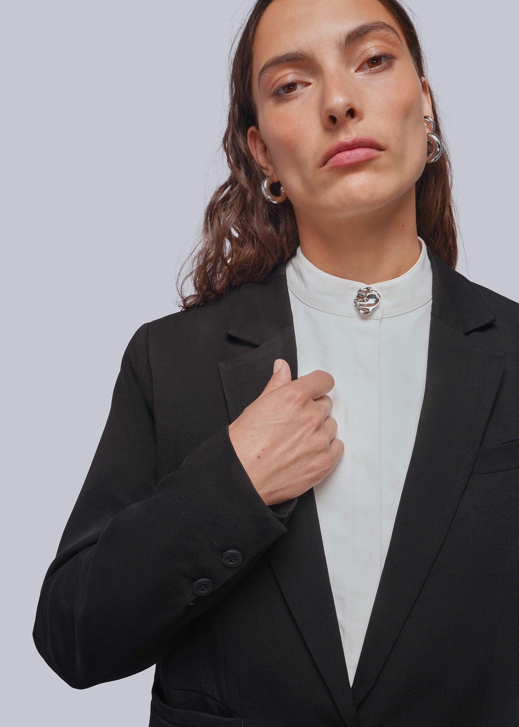 A woman wearing a black blazer over a white mock-neck top with a silver decorative button cover at collar, set against a solid light gray background.