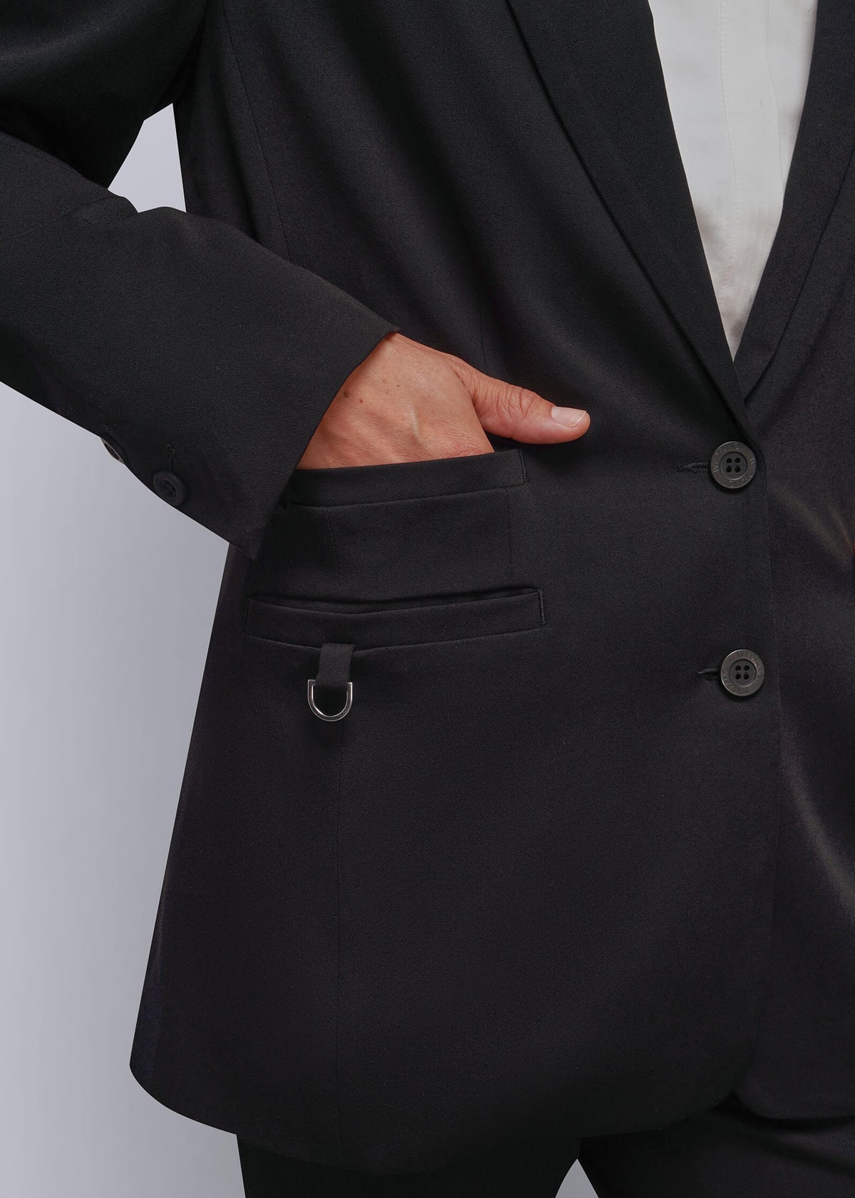 S=Close-up of a person wearing a black tailored blazer with a notched lapel, two front buttons, a pocket with a d-ring detail, and matching black trousers against a light gray studio background.