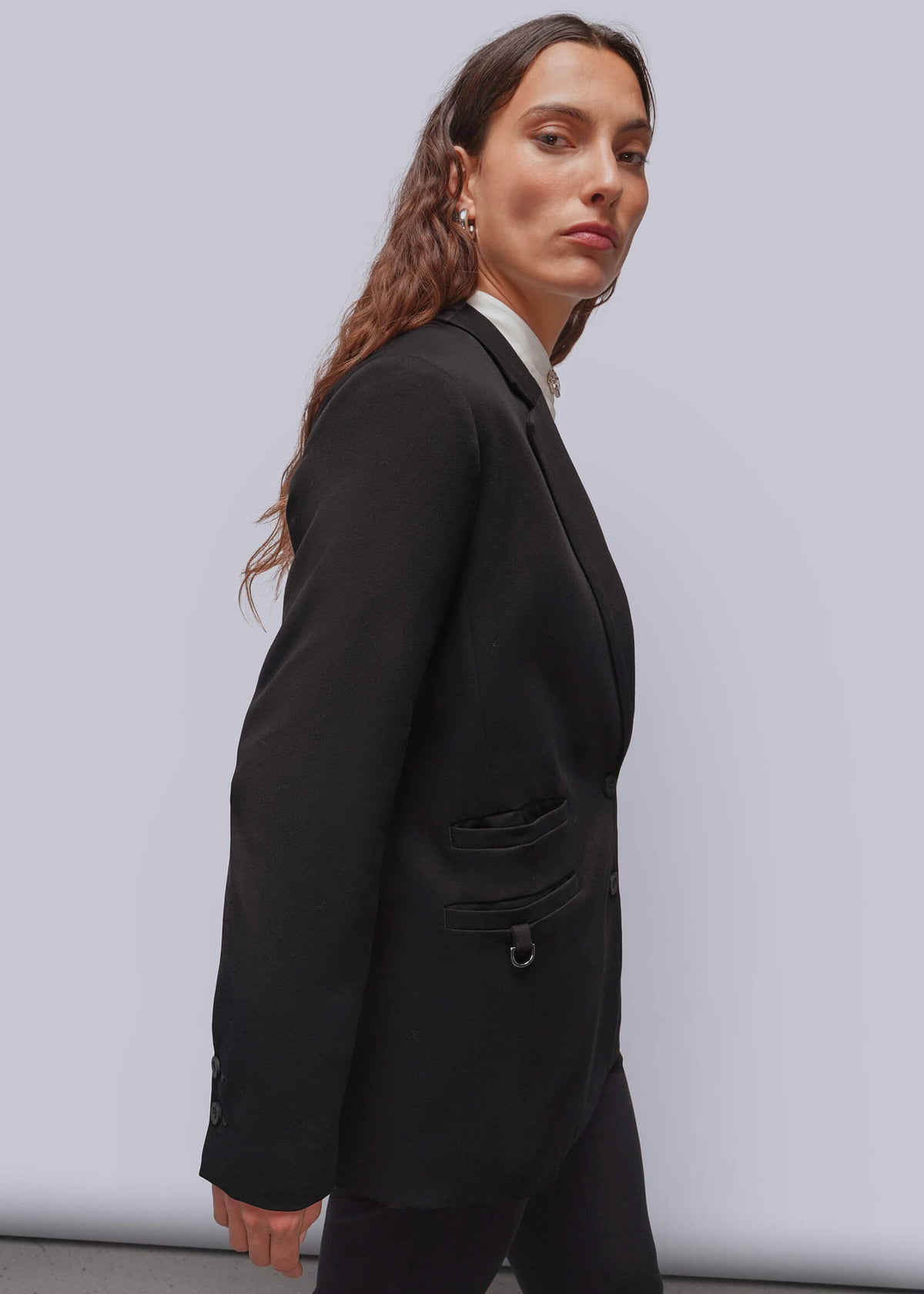 S=A woman wearing a black tailored blazer over a white collared shirt, shown in side profile against a plain light gray studio background.