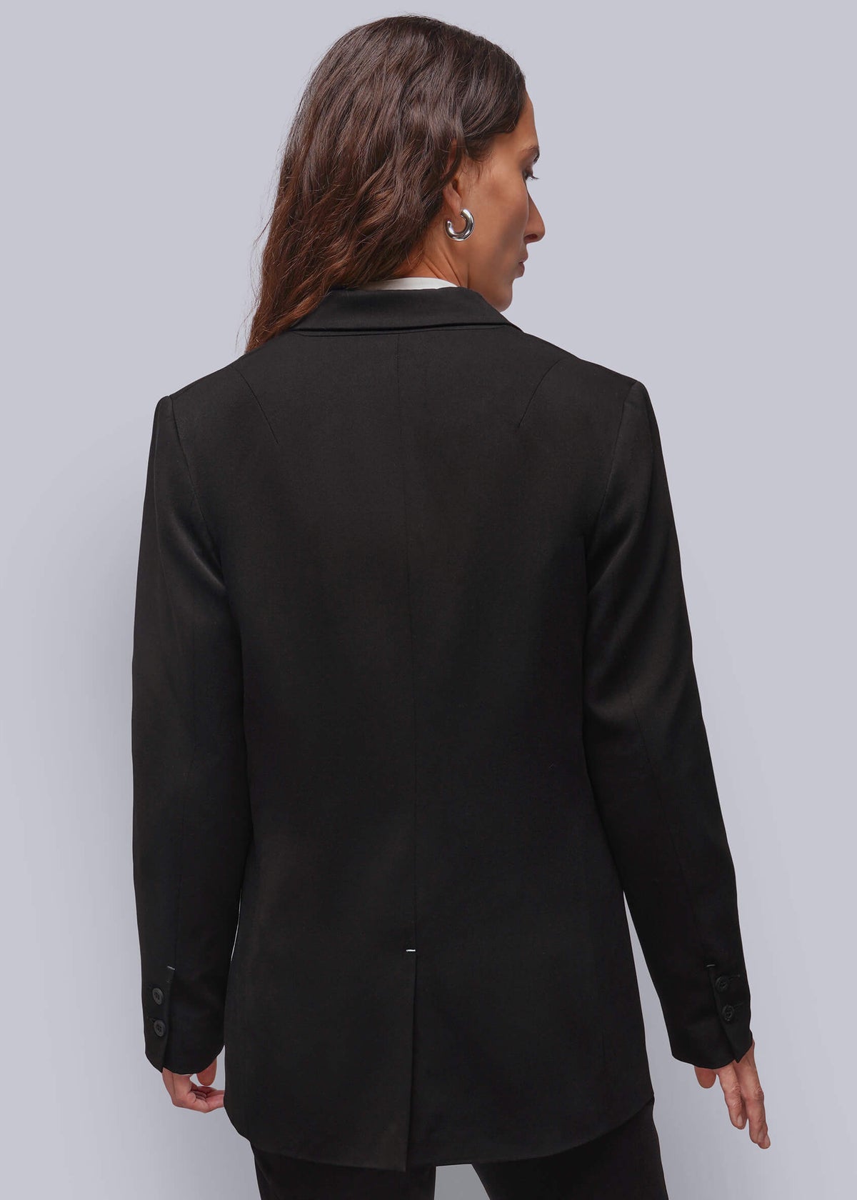 S=A woman shown from the back wearing a black blazer over a white collared shirt, paired with black trousers, against a solid light gray studio background.